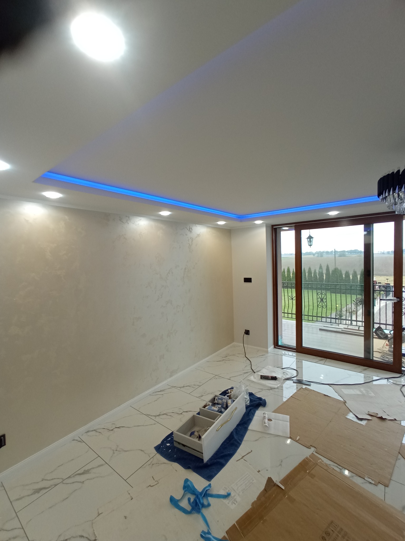 Wykończone wnętrze pokoju z dekoracyjną ścianą w odcieniach beżu, podświetleniem LED na suficie i marmurową podłogą. Widoczne narzędzia i materiały malarskie.