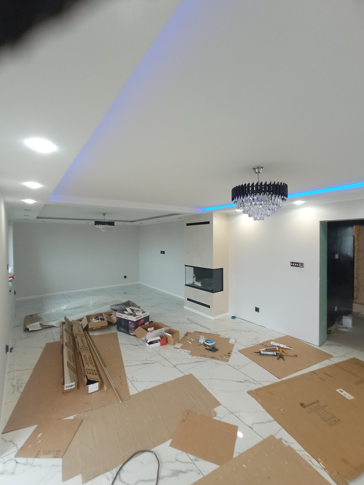 Jasne wnętrze z marmurową podłogą i nowoczesnym kominkiem. Sufit z oświetleniem LED. Na podłodze kartony i narzędzia, sugerujące prace wykończeniowe w toku.