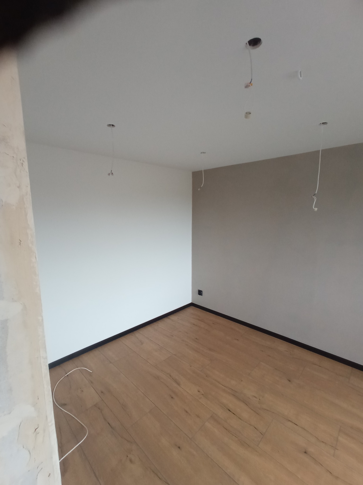 Pokój po remoncie: drewniana podłoga, pomalowane ściany w odcieniach bieli i beżu, widoczne kable oświetleniowe. Fragment ściany w stanie surowym po lewej.
