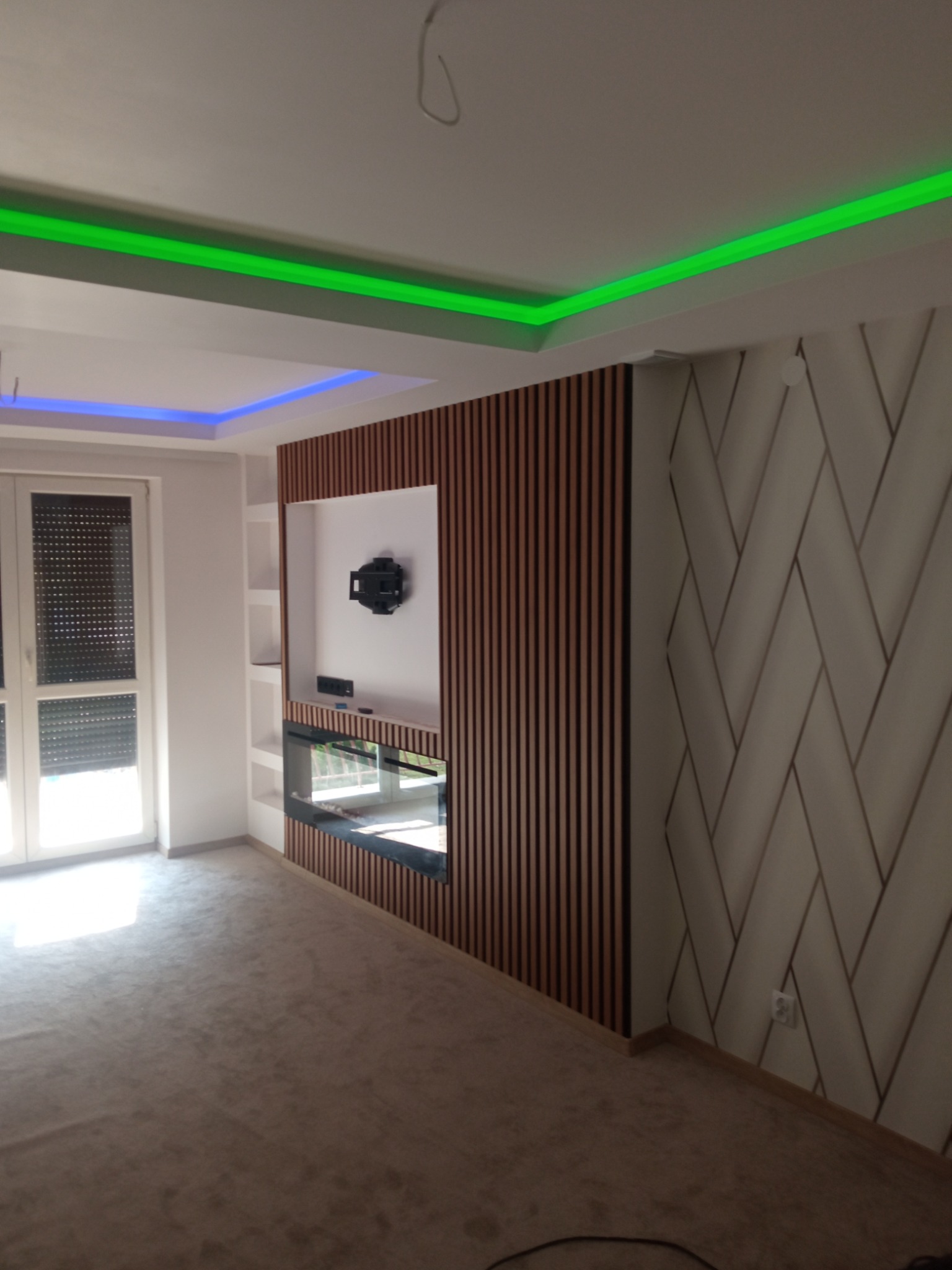 Nowoczesne wykończenie wnętrza: ściana z drewnianych lameli z wnęką na TV i kominek, dekoracyjna ściana z geometrycznym wzorem, podświetlany sufit LED (zielony i niebieski).