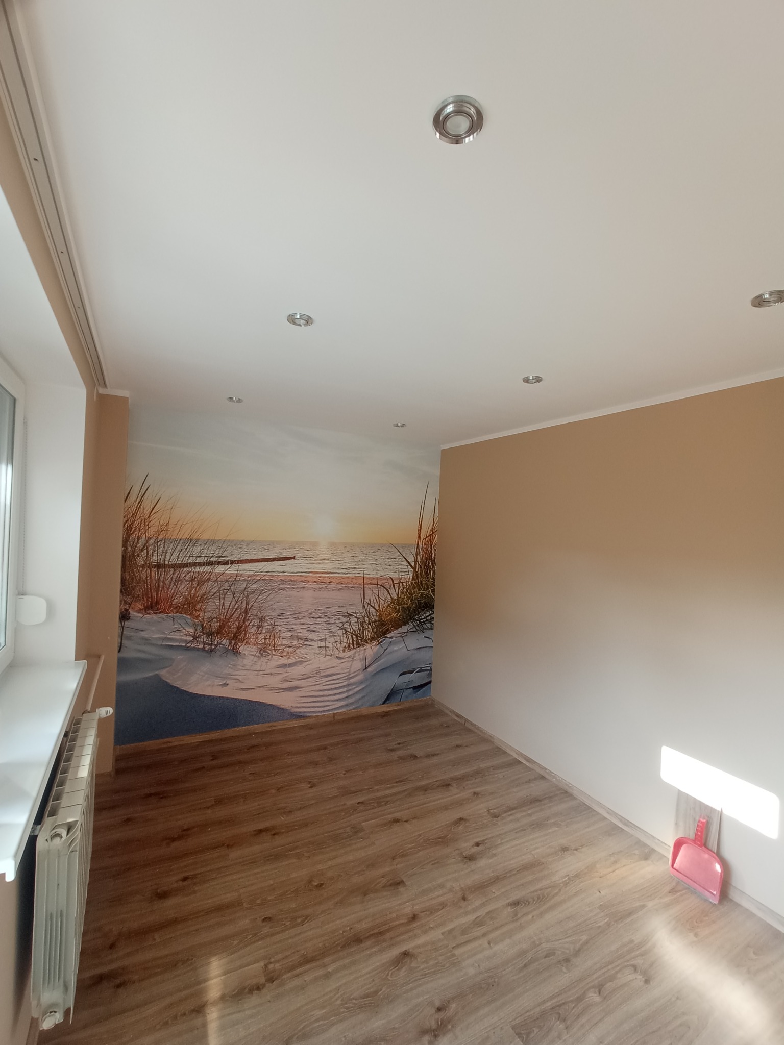 Pokój po remoncie z fototapetą plaży i pomalowanymi ścianami na beżowo. Na podłodze panele, widoczny grzejnik i różowa szufelka.