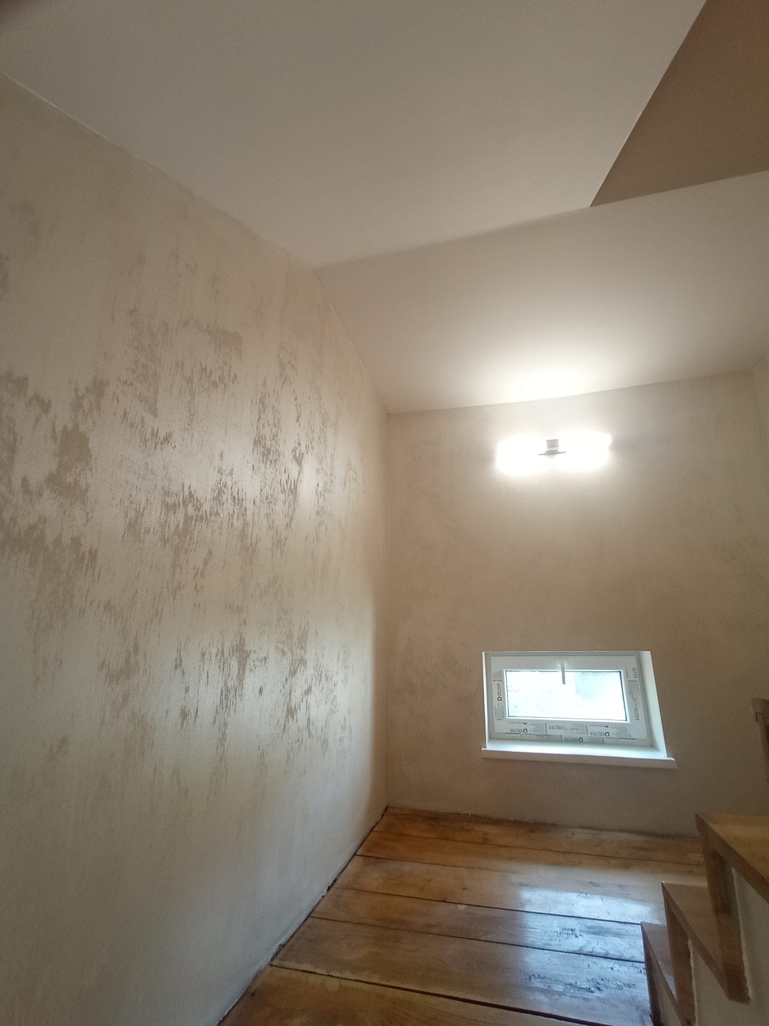 Ściana z dekoracyjną, beżową strukturą, małe okno z białymi ramami, drewniana podłoga i schody. Oświetlenie sufitowe.