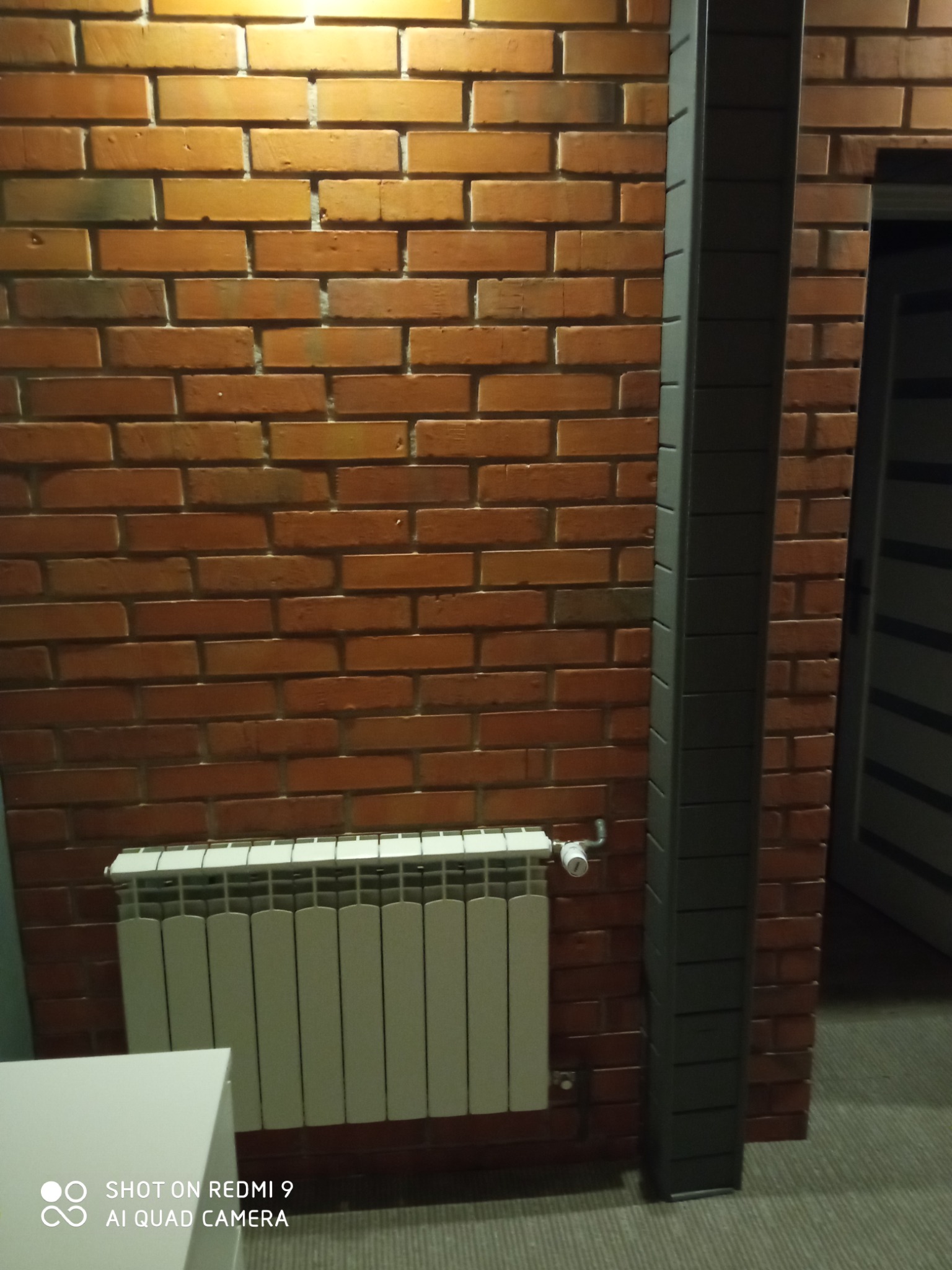 Ceglana ściana z grzejnikiem i szarą kolumną dekoracyjną. Widoczny fragment białego mebla i drzwi w tle. Ciepłe oświetlenie.