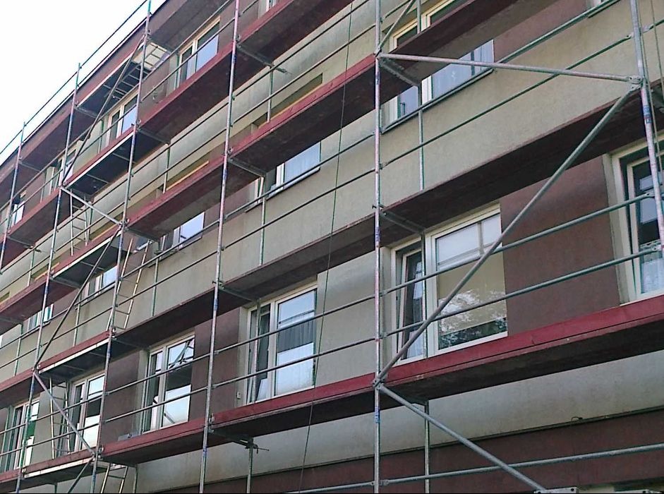 Budynek wielorodzinny w trakcie prac elewacyjnych. Widoczne rusztowania i fragmenty ocieplonej oraz pomalowanej ściany w odcieniach szarości i brązu.