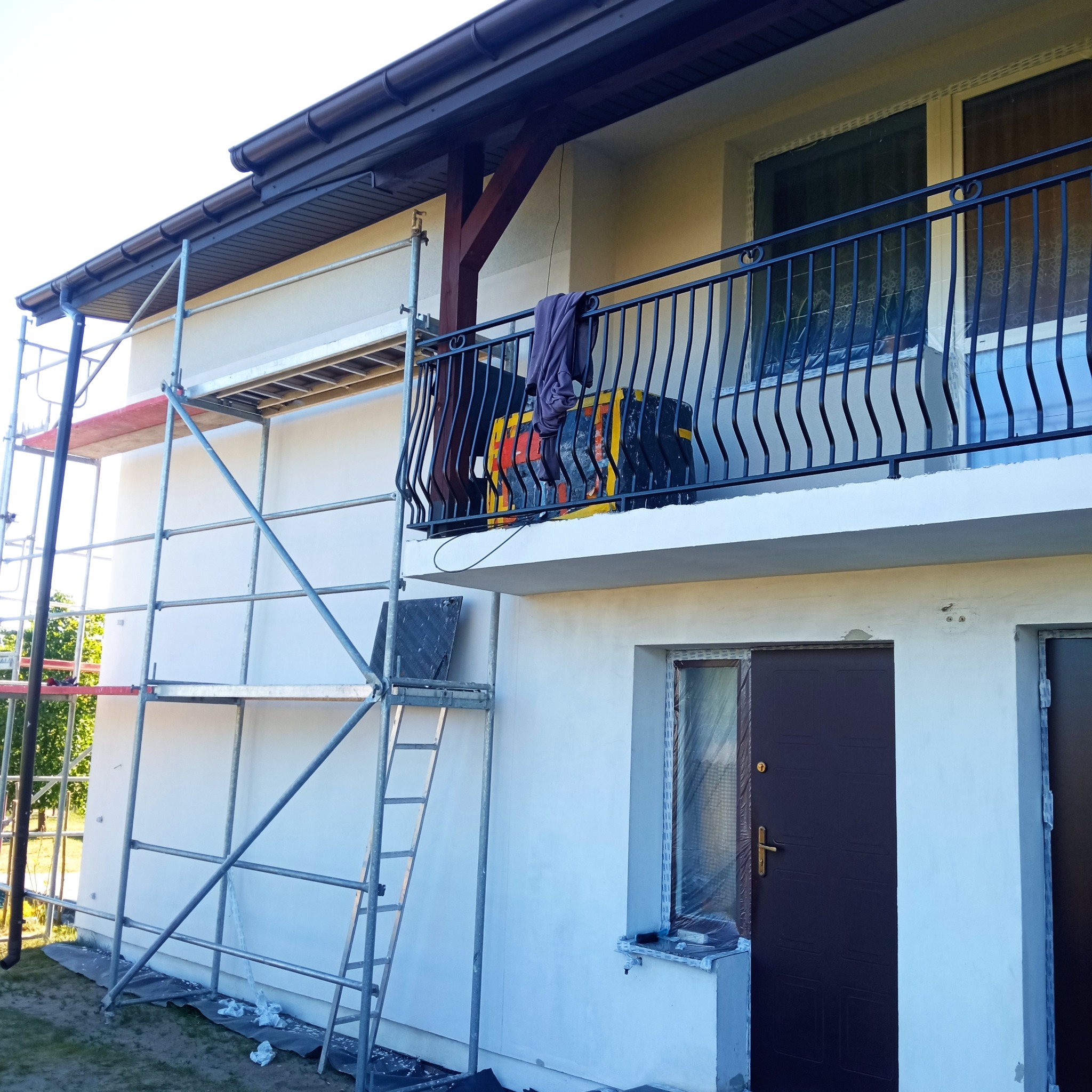 Dom z rusztowaniem podczas prac elewacyjnych. Widoczny balkon z czarną balustradą, okna zabezpieczone folią i świeżo pomalowana, jasna elewacja.