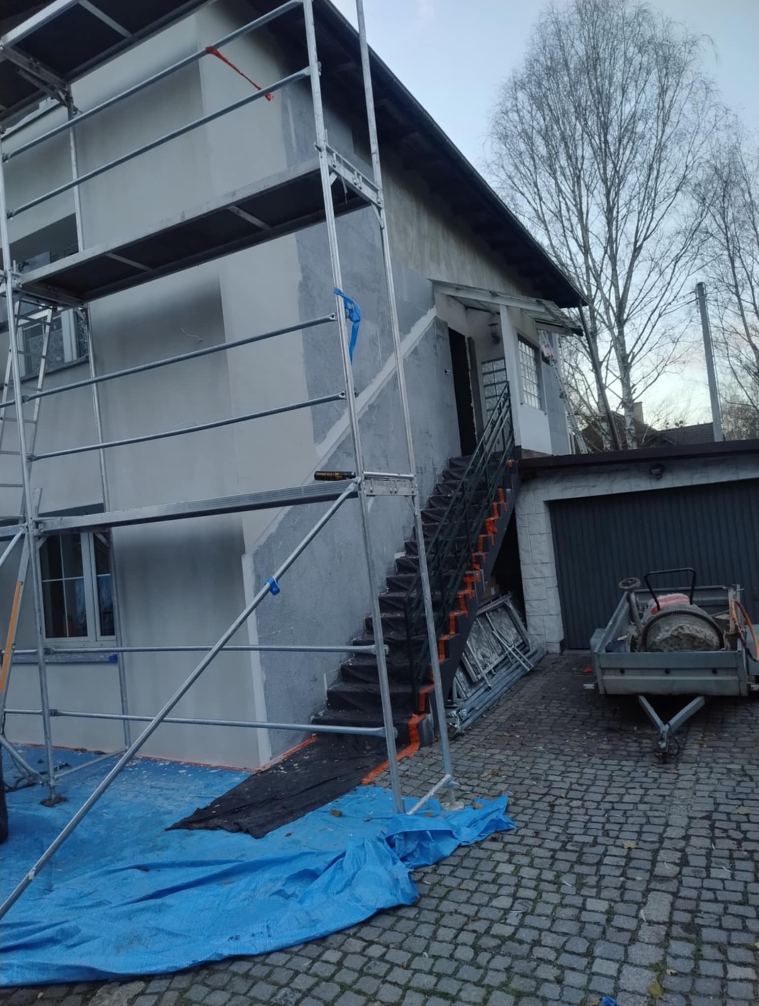 Dom w trakcie prac elewacyjnych: szare ściany, rusztowania, zabezpieczenia. Widoczne schody zewnętrzne i przyczepka z betoniarką. Prace w toku, bez wykończenia.