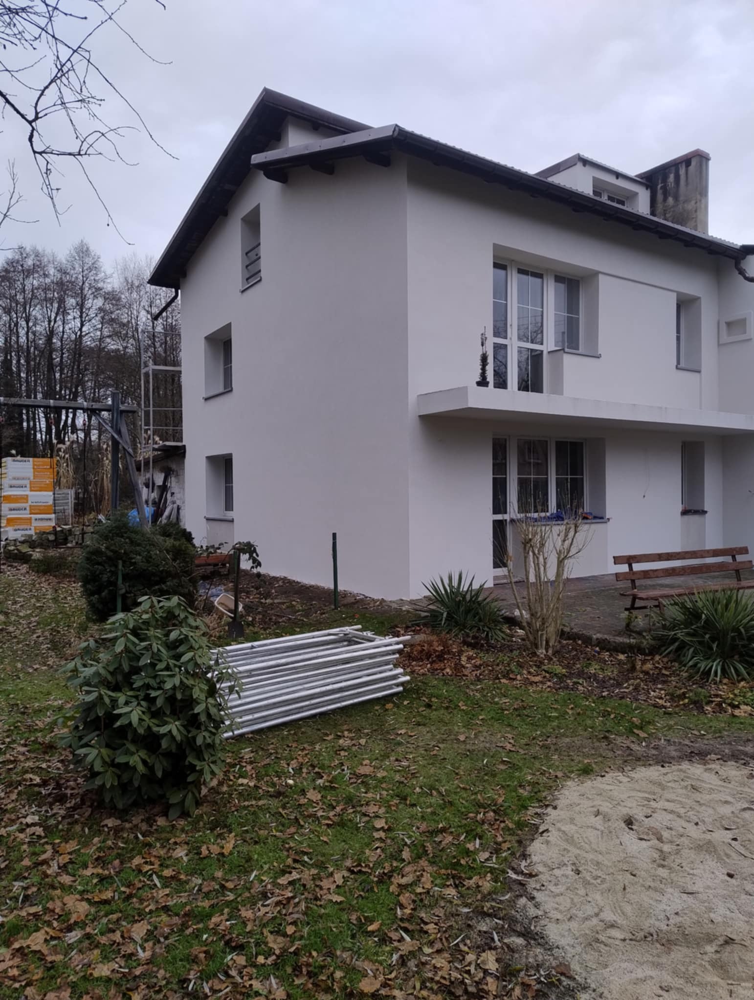 Biały, dwupiętrowy dom po renowacji elewacji, z widocznymi rusztowaniami i stertą metalowych elementów na trawniku. Świeżo pomalowana fasada w jesiennej scenerii.