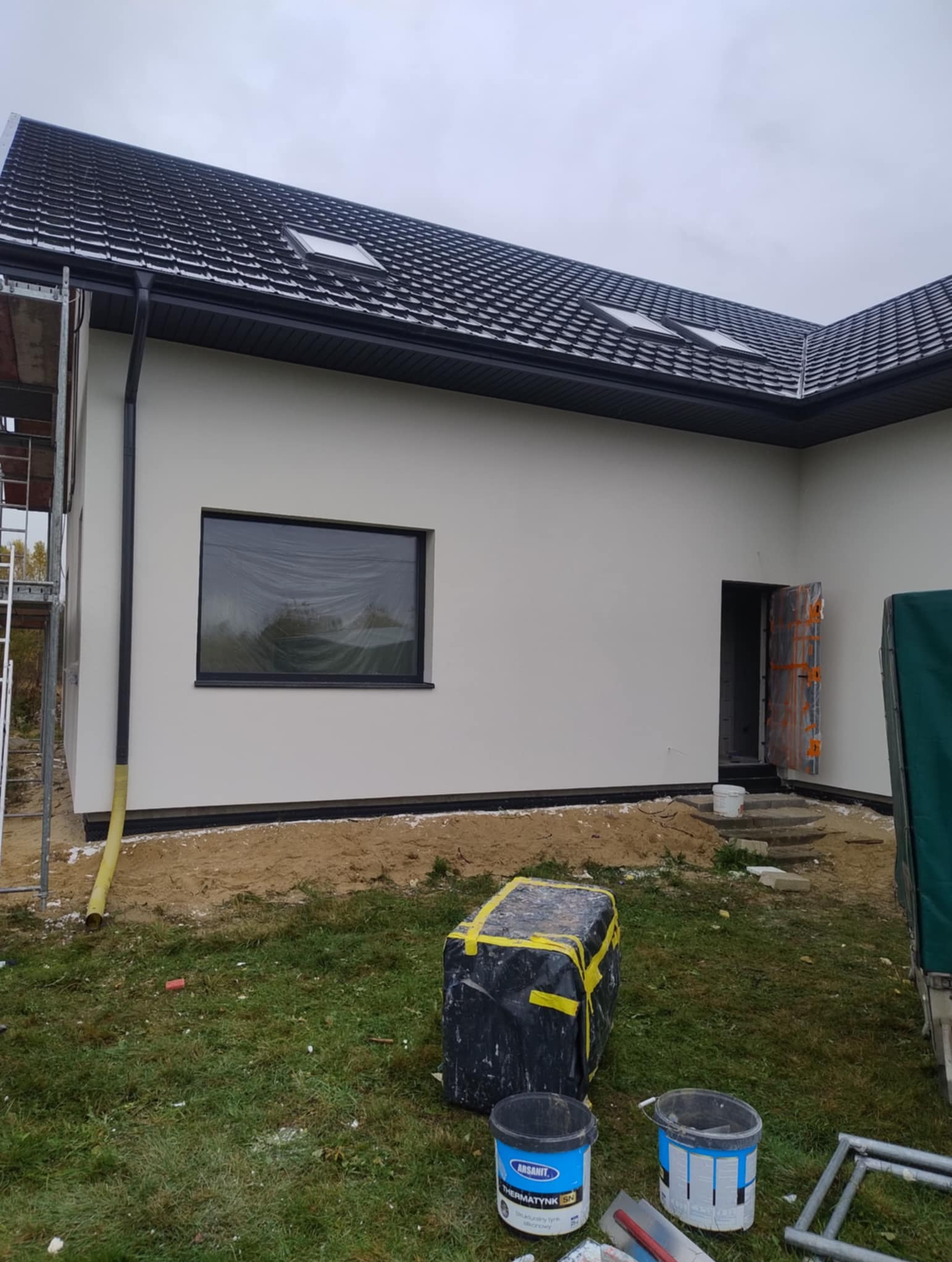 Jasna elewacja domu jednorodzinnego z czarną dachówką i oknami. Na pierwszym planie wiadro z farbą i narzędzia. Częściowo widoczne rusztowanie.