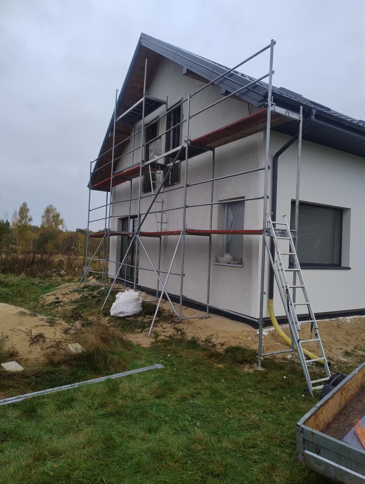 Dom jednorodzinny w trakcie prac elewacyjnych. Widoczne rusztowania, ściany pokryte szarą warstwą, okna zabezpieczone folią. Na trawie leżą elementy rusztowania.