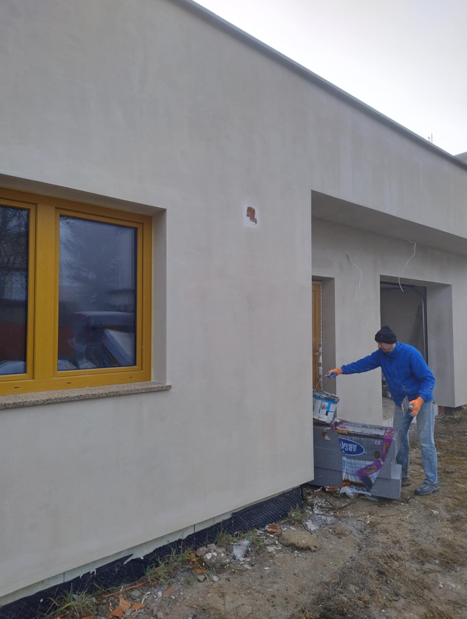 Pracownik w trakcie malowania elewacji domu jednorodzinnego na szaro. Widoczne okno z żółtą ramą, puszka z farbą i narzędzia. Fragment ocieplenia przy ziemi.
