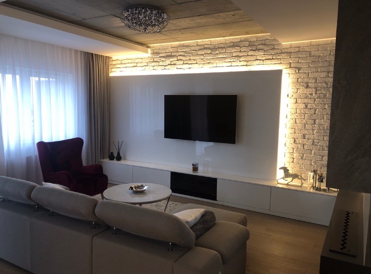 Nowoczesny salon z białymi szafkami RTV pod telewizorem, ceglaną ścianą z podświetleniem LED, szarym narożnikiem i bordowym fotelem.