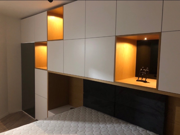 Biało-drewniana szafa wnękowa z podświetlanymi półkami nad łóżkiem, wykonana na wymiar w Rzeszowie, z dekoracyjną figurką w jednej z wnęk.