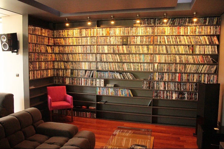 Ogromna, ciemnoszara biblioteczka na całą ścianę, wypełniona płytami CD i DVD, z czerwonym fotelem po lewej stronie i brązową sofą na pierwszym planie.