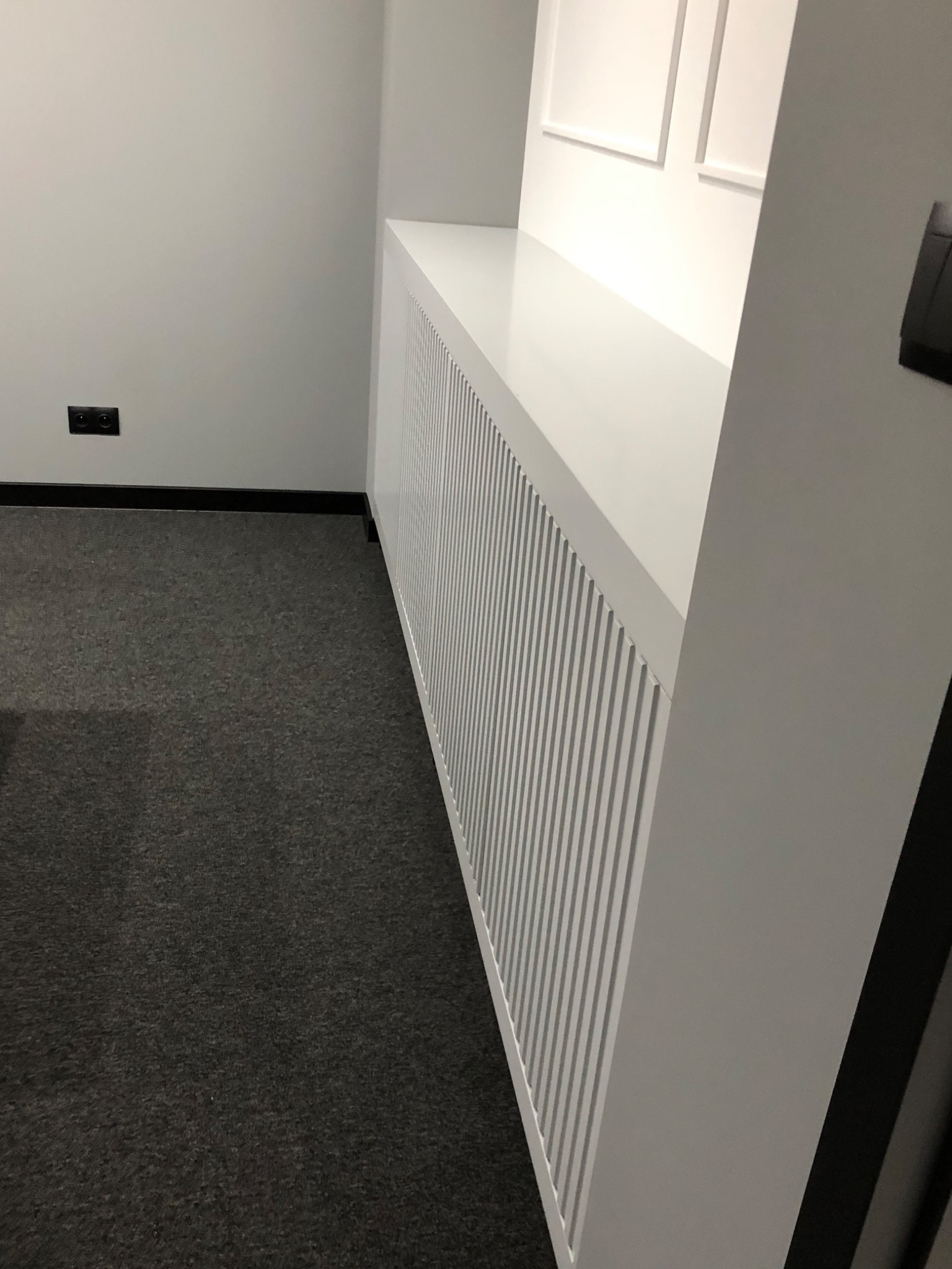 Biała zabudowa grzejnika z pionowymi listwami i blatem, dopasowana do wnętrza z szarą wykładziną i białymi ścianami.