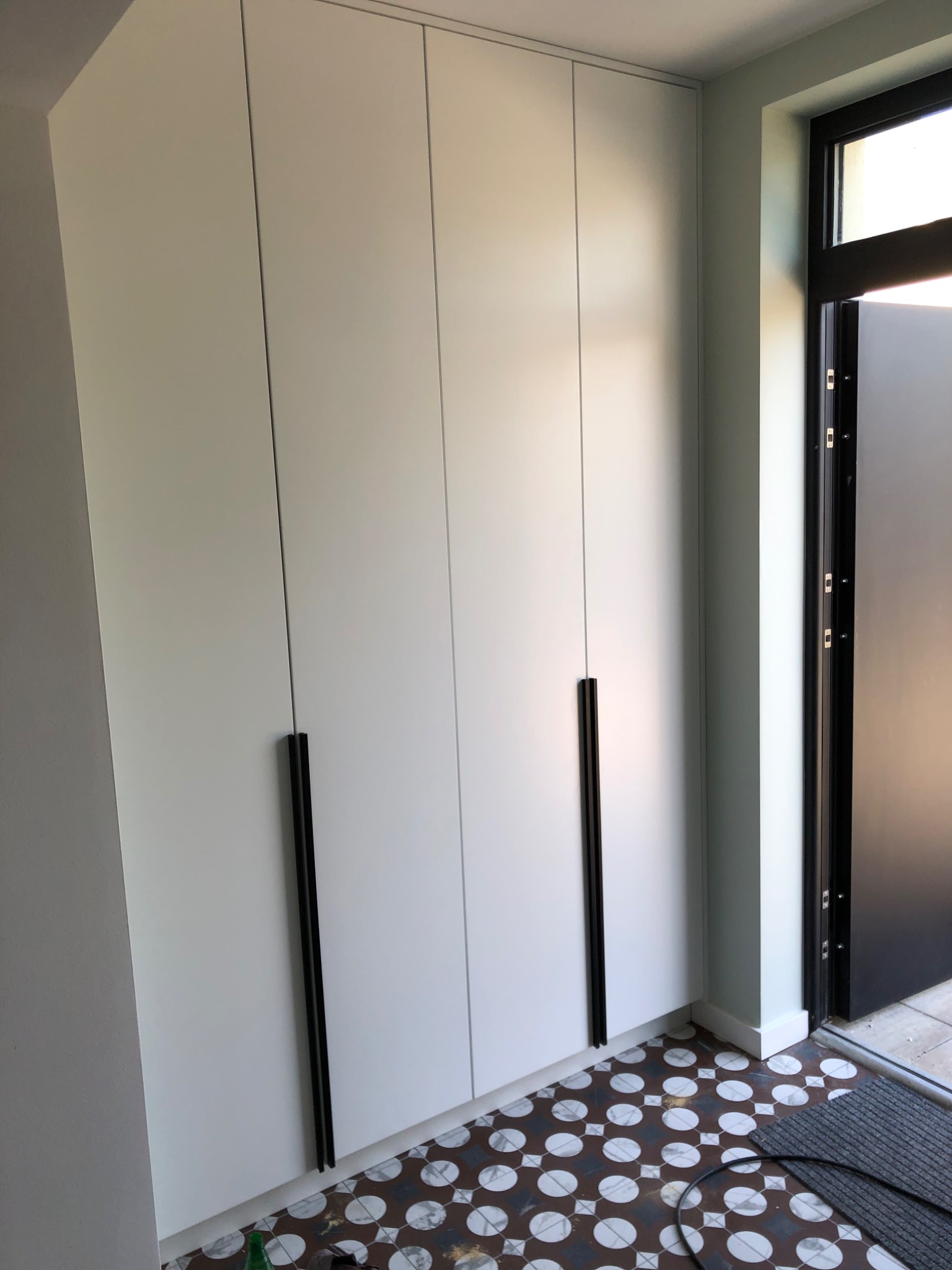 Biała szafa wnękowa z długimi, czarnymi uchwytami, widoczna przy wejściu z charakterystyczną, wzorzystą podłogą.