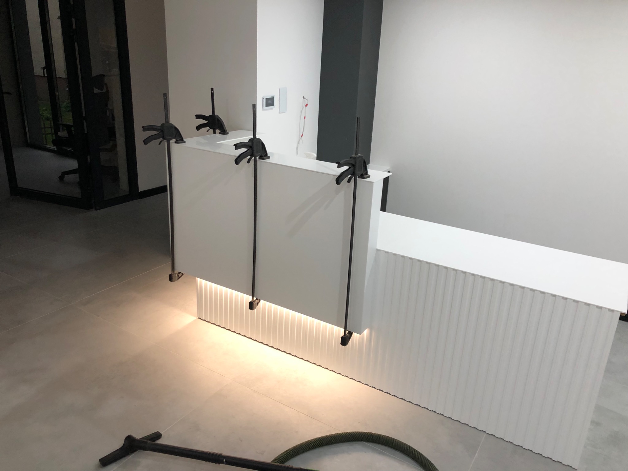 Biała lada recepcyjna w trakcie montażu, zabezpieczona ściskami stolarskimi, widoczny wąż odkurzacza na podłodze.