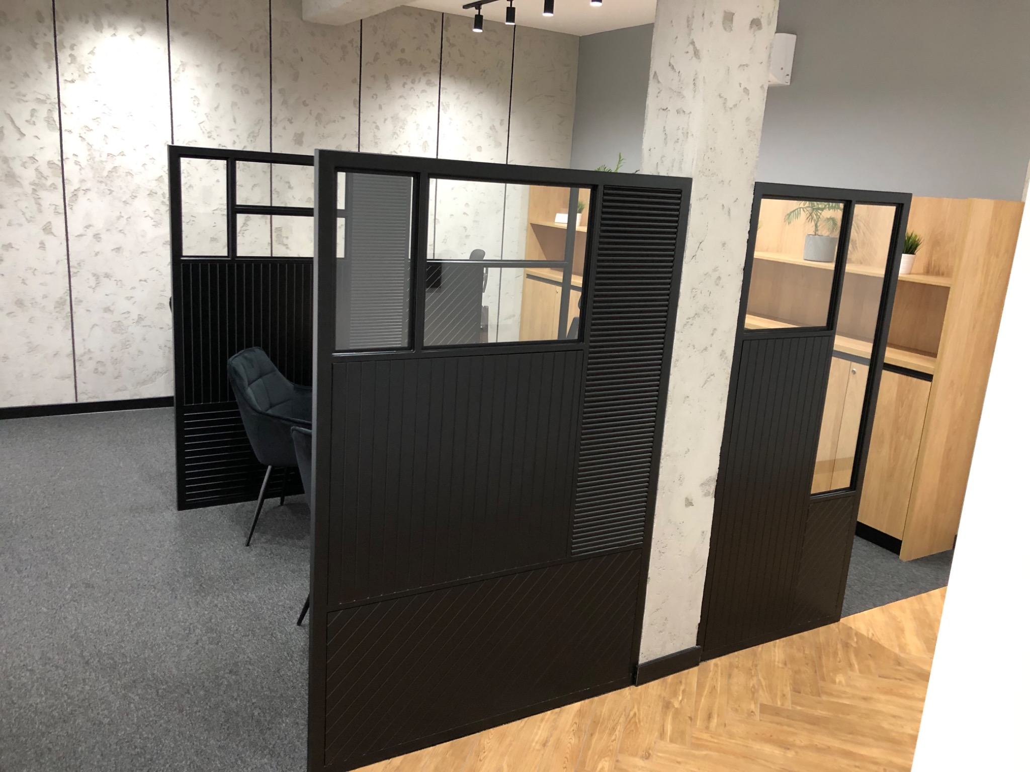 Nowoczesne, czarne ścianki działowe z przeszkleniami i żaluzjami w biurze z szarą wykładziną i parkietem w jodełkę.
