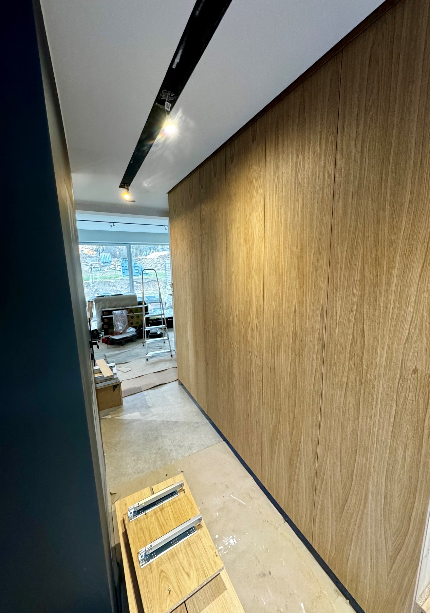 Długa, drewniana szafa wnękowa z jasnego drewna, zamontowana w korytarzu podczas prac wykończeniowych, widoczne elementy montażowe i drabina w tle.