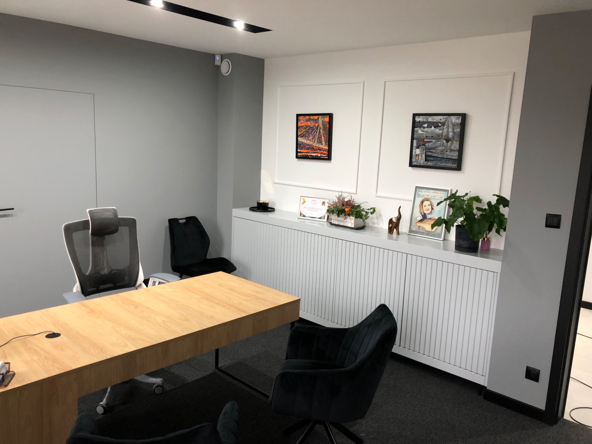 Jasny gabinet z drewnianym biurkiem, szarymi ścianami i białymi szafkami z pionowymi żłobieniami, dekorowany obrazami i roślinami doniczkowymi.