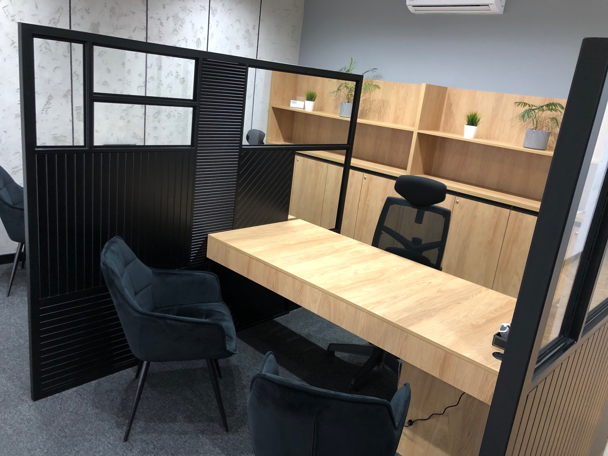 Nowoczesne biuro z drewnianym biurkiem, szafkami i czarną ażurową ścianką działową oraz krzesłami z ciemnego weluru.