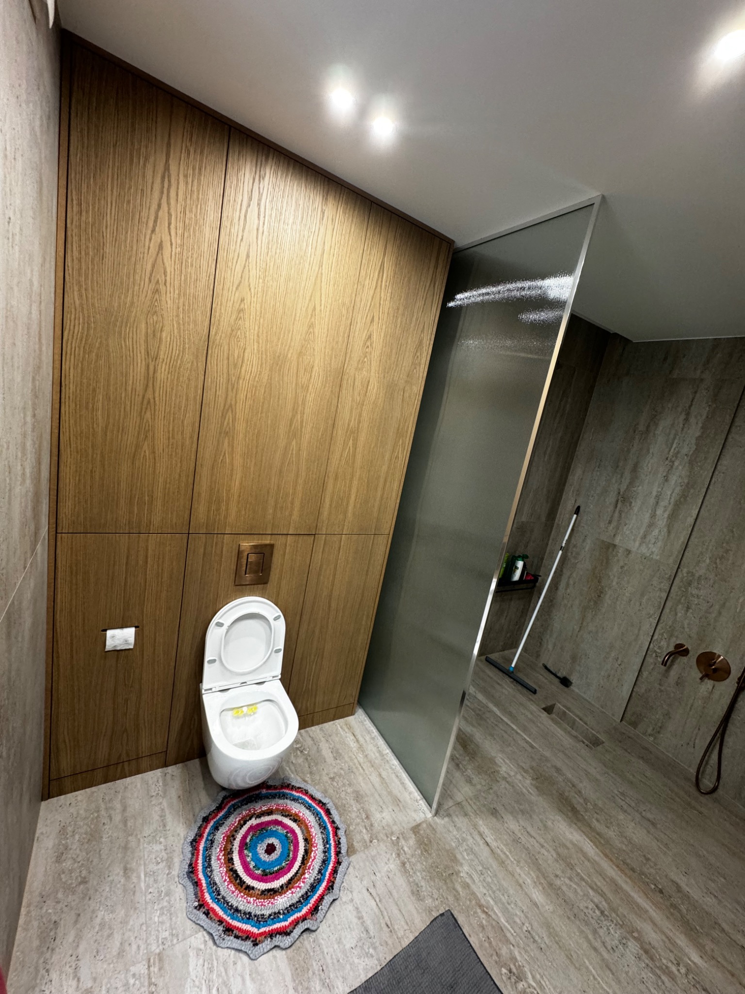 Nowoczesna łazienka z drewnianą zabudową, toaletą i szklaną kabiną prysznicową, akcentowana kolorowym dywanikiem pod toaletą.