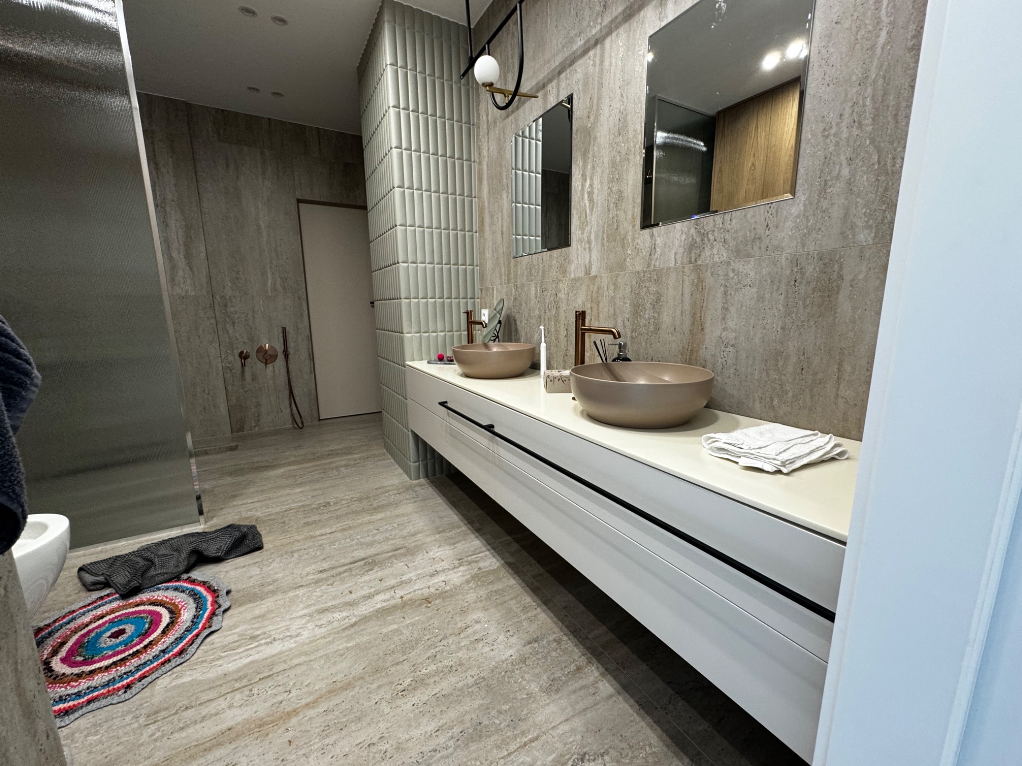 Nowoczesna łazienka z dwoma okrągłymi umywalkami w kolorze taupe na białym blacie z czarnymi uchwytami, jasne płytki imitujące kamień na ścianach, dekoracyjna lampa sufitowa i dwa lustra.