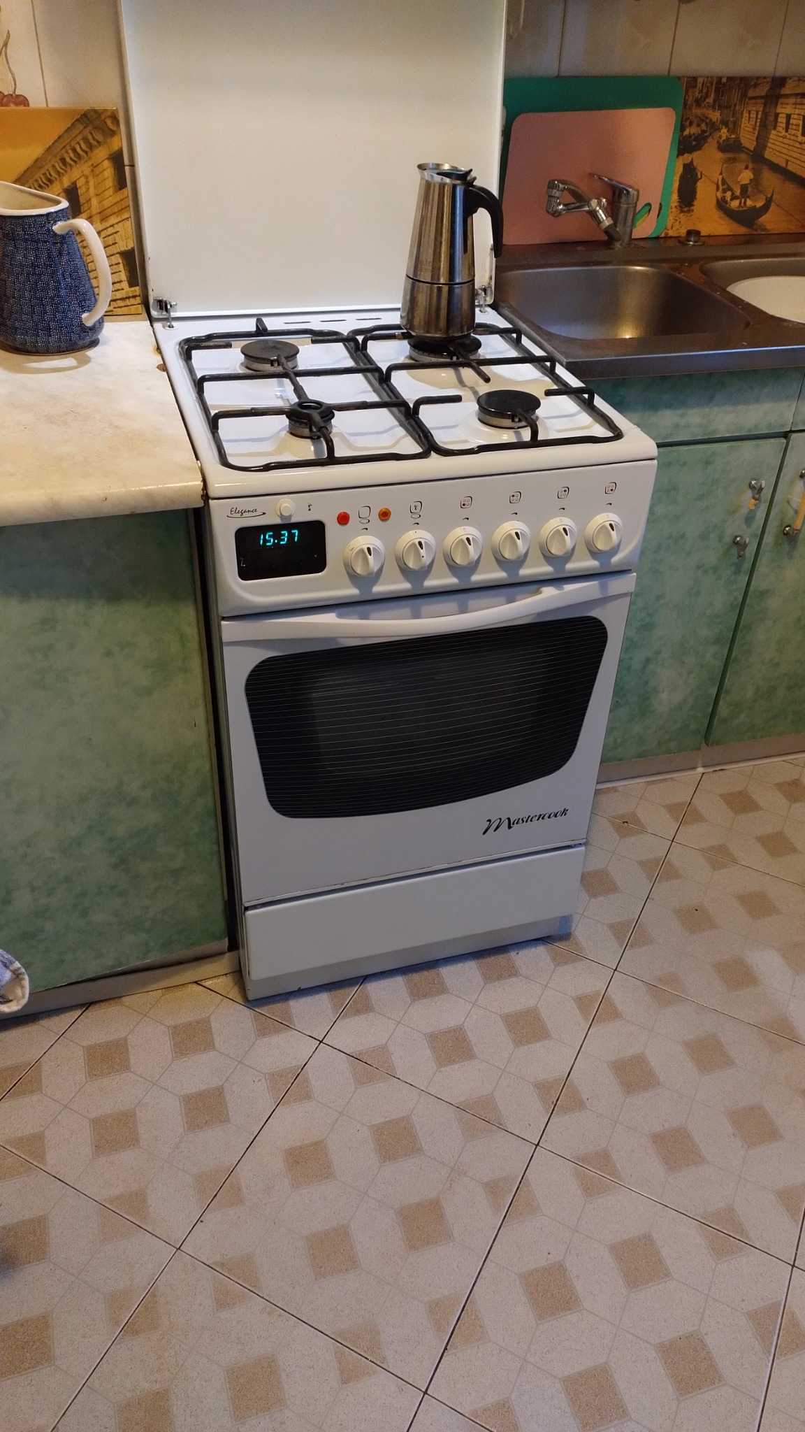 Biała kuchenka gazowa Mastercook z czarnymi rusztami i wyświetlaczem zegara, na której stoi srebrna kawiarka.