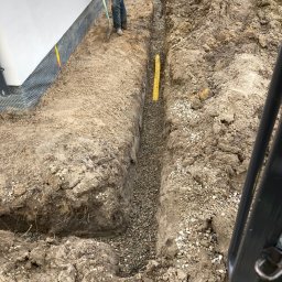 ROL-KOP - Wykop z żółtą rurą i drenażem z kamieni, pracownik z łopatą przy ścianie budynku z folią kubełkową. Prace ziemne w trakcie realizacji.
