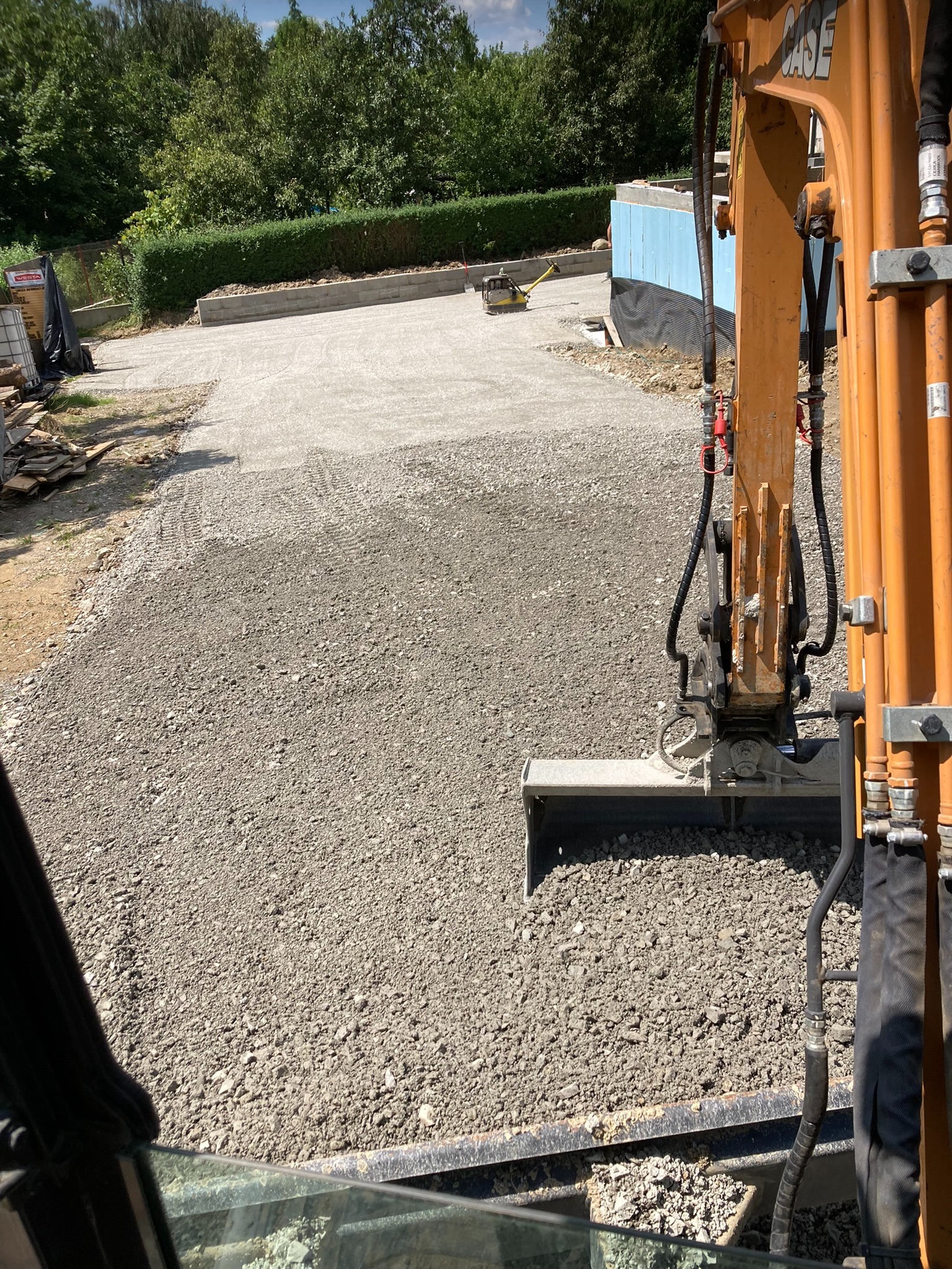 Utwardzony teren z kruszywem, widok z kabiny koparki, w tle zagęszczarka i zielona roślinność. Widoczne ślady pracy maszyn.