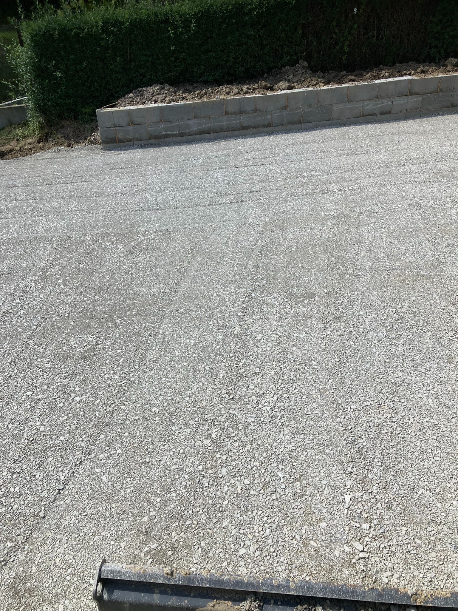 Utwardzona droga z szarego kruszywa, obrzeża z betonowych bloczków. Widok z góry, z dołu fragment maszyny budowlanej. Zielony żywopłot w tle.