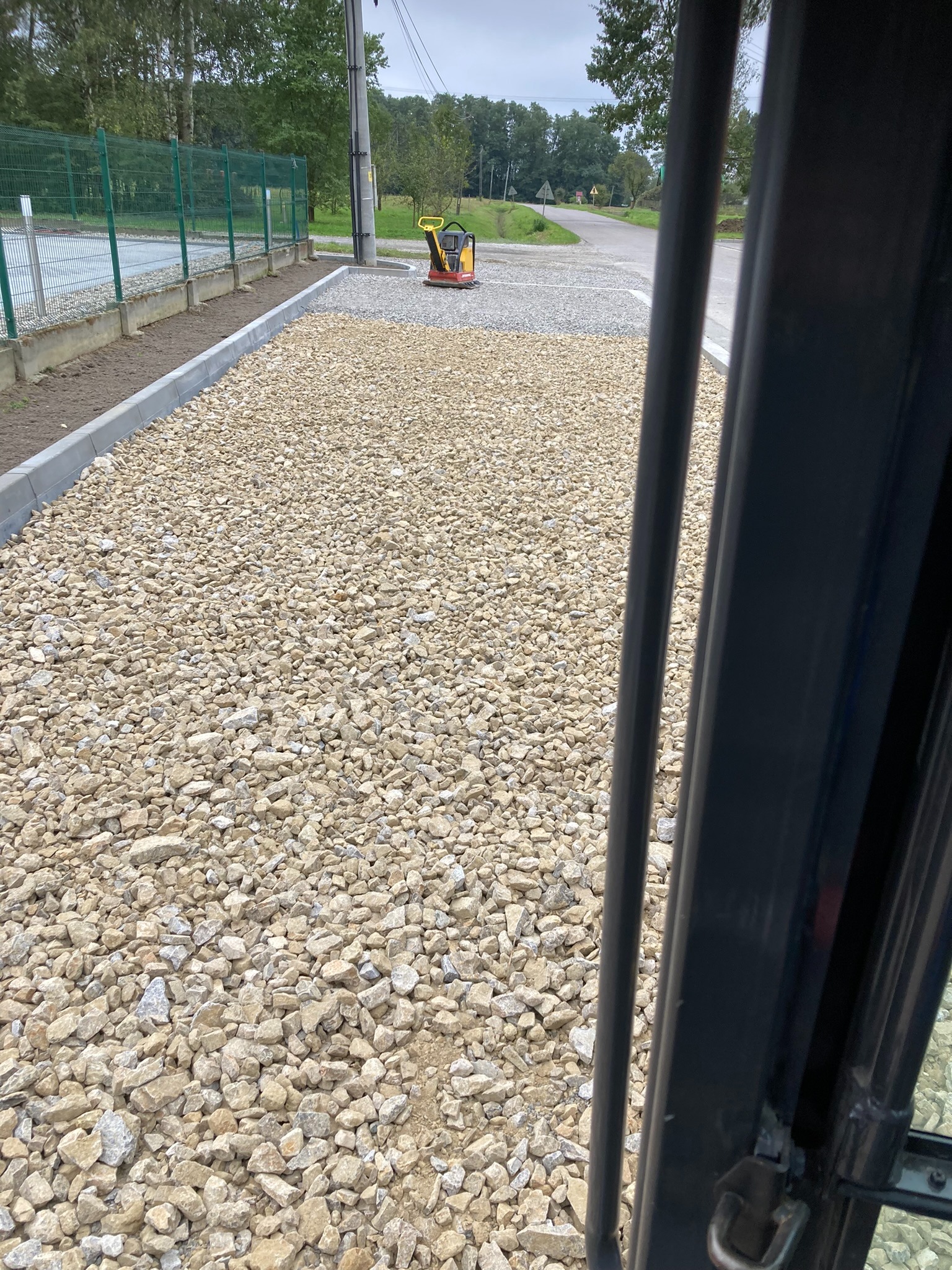 Utwardzona droga z kruszywa, widok z kabiny maszyny budowlanej, na tle zieleni i ogrodzenia. W tle zagęszczarka wibracyjna.