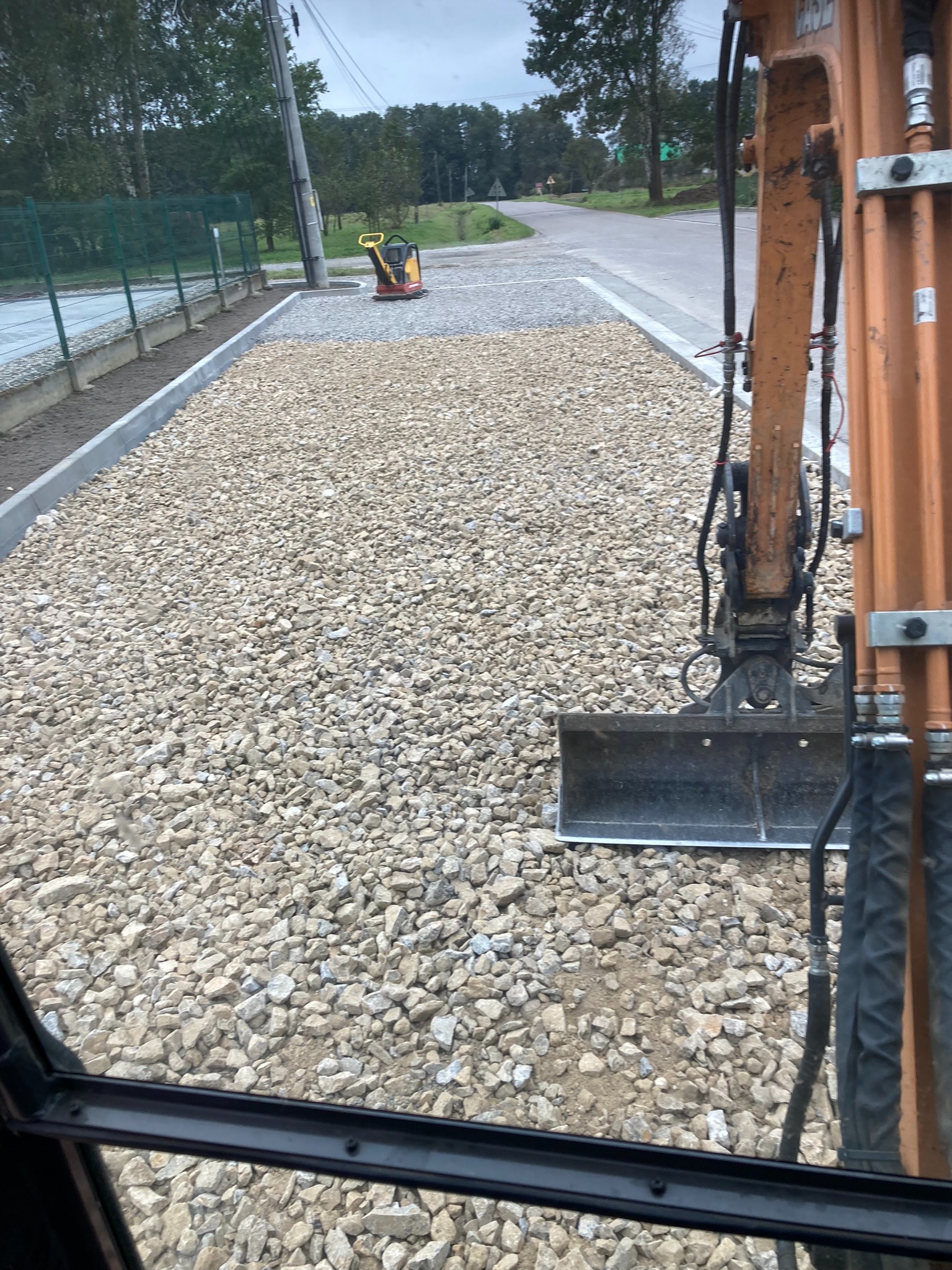 Utwardzony teren z użyciem żwiru, widok z kabiny koparki, mała zagęszczarka w tle. Obrzeża chodnika wykończone. Prace drogowe w toku, widok na ulicę i zieleń.