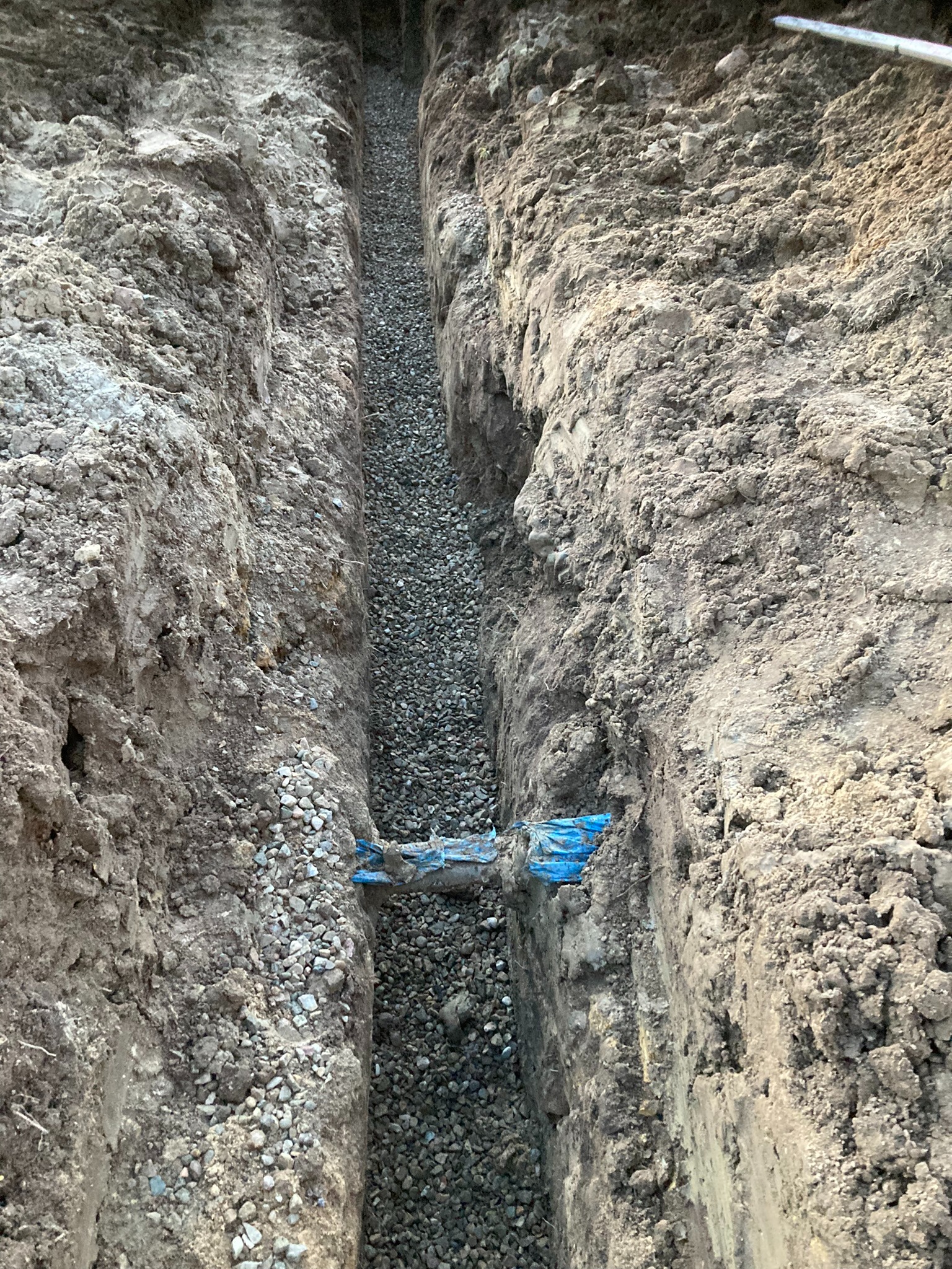Wykopany wąski rów wypełniony drobnym żwirem, z widocznym fragmentem niebieskiej rury. Ściany rowu z ubitej ziemi, nierówne. Ujęcie z góry.