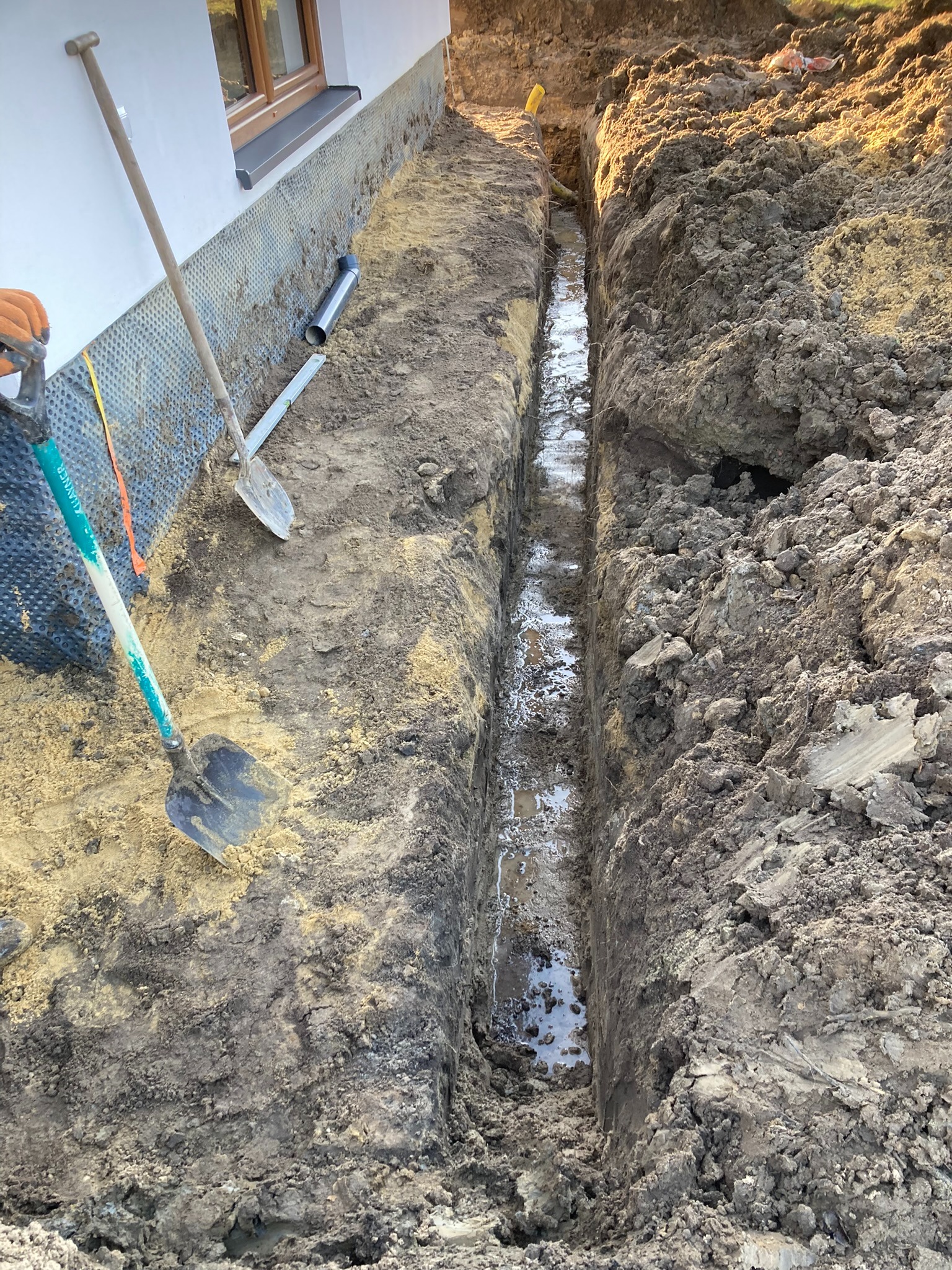 Wykopany wąski rów wzdłuż fundamentów budynku, z widoczną folią kubełkową i fragmentem rury. W tle sterta ziemi i fragment żółtej rury. Dwie łopaty oparte o ścianę.