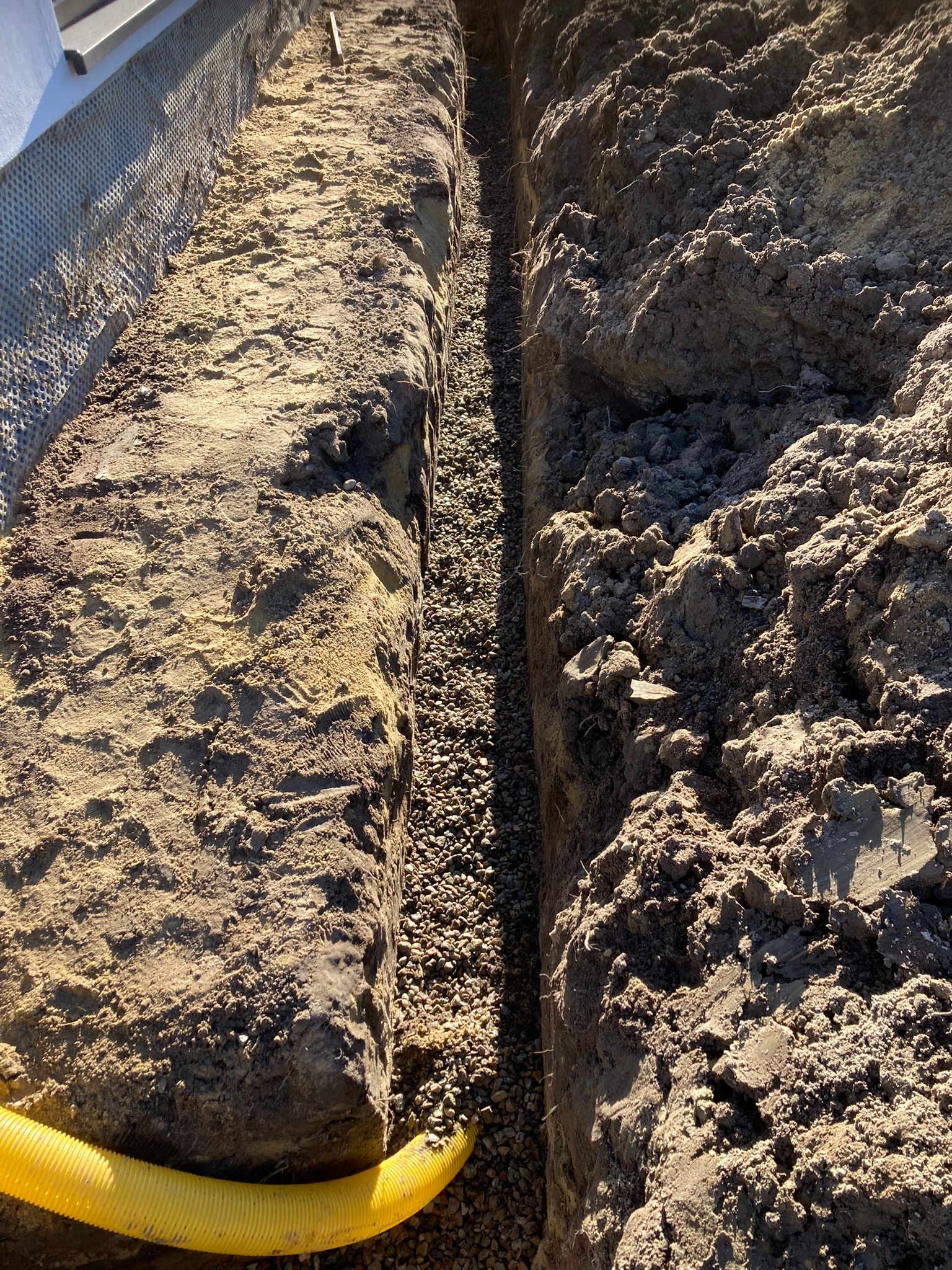 Wykop pod fundament z drenażem: wąski rów wypełniony żwirem, żółta rura drenażowa na dnie, widoczna izolacja pionowa budynku. Gotowy etap prac ziemnych.