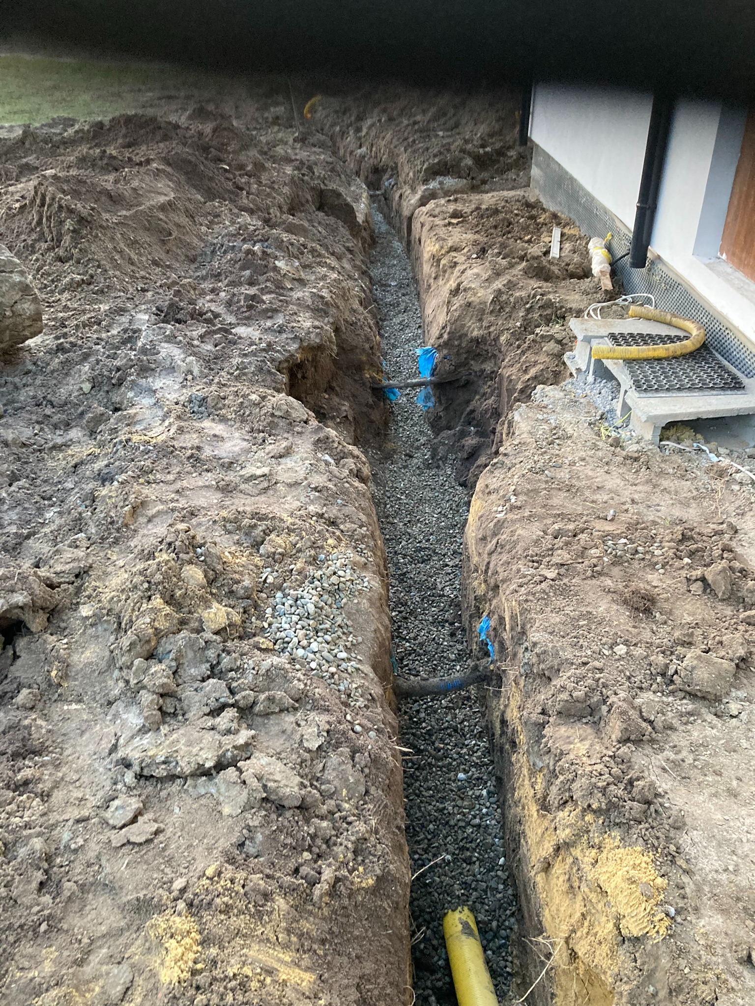 Wykopany rów z ułożonym drenażem z rur i kamieni, widoczny fragment budynku z izolacją. Wykop ziemny z żółtymi rurami drenażowymi i niebieskimi elementami.