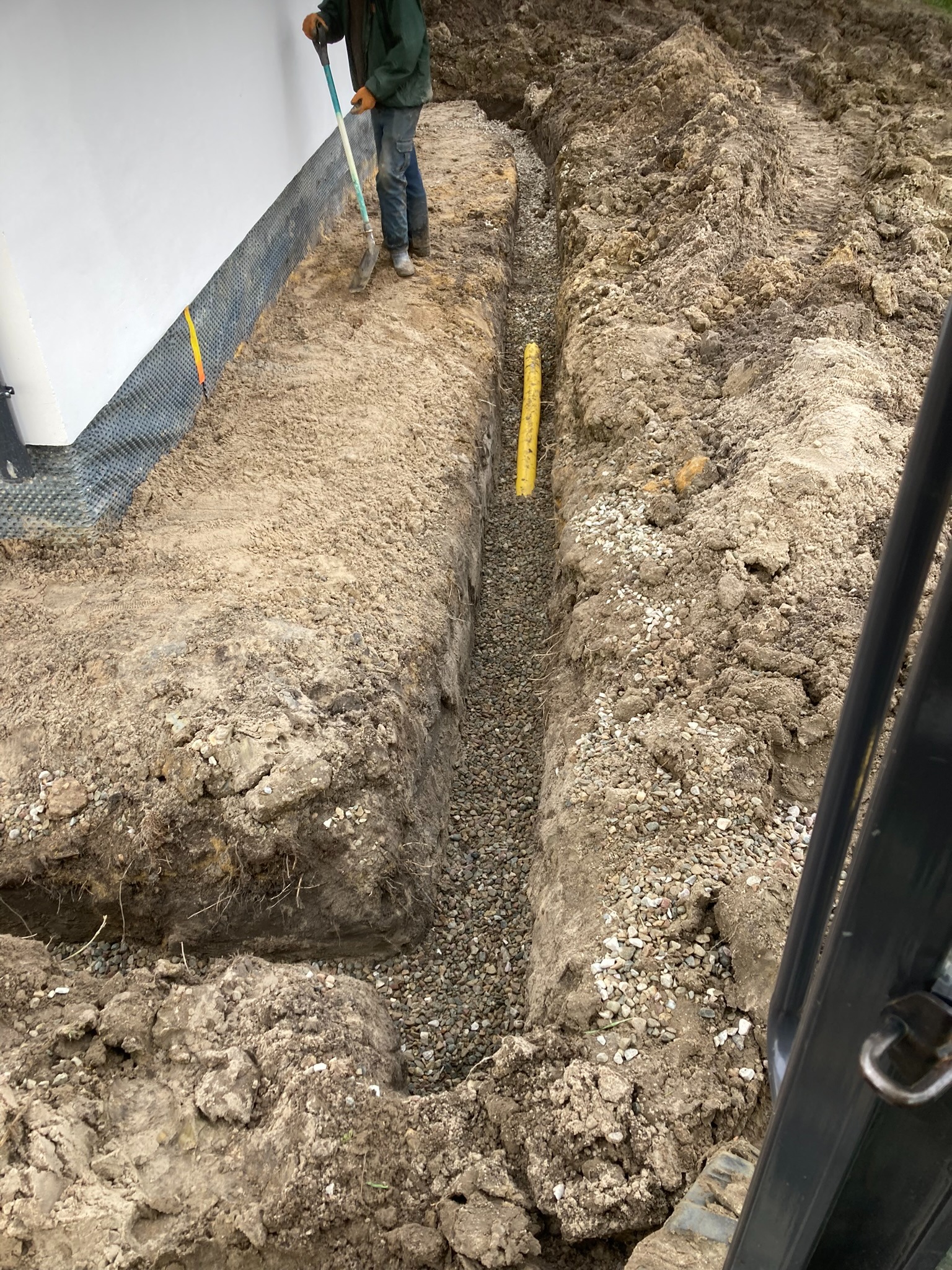 Wykop z żółtą rurą i drenażem z kamieni, pracownik z łopatą przy ścianie budynku z folią kubełkową. Prace ziemne w trakcie realizacji.