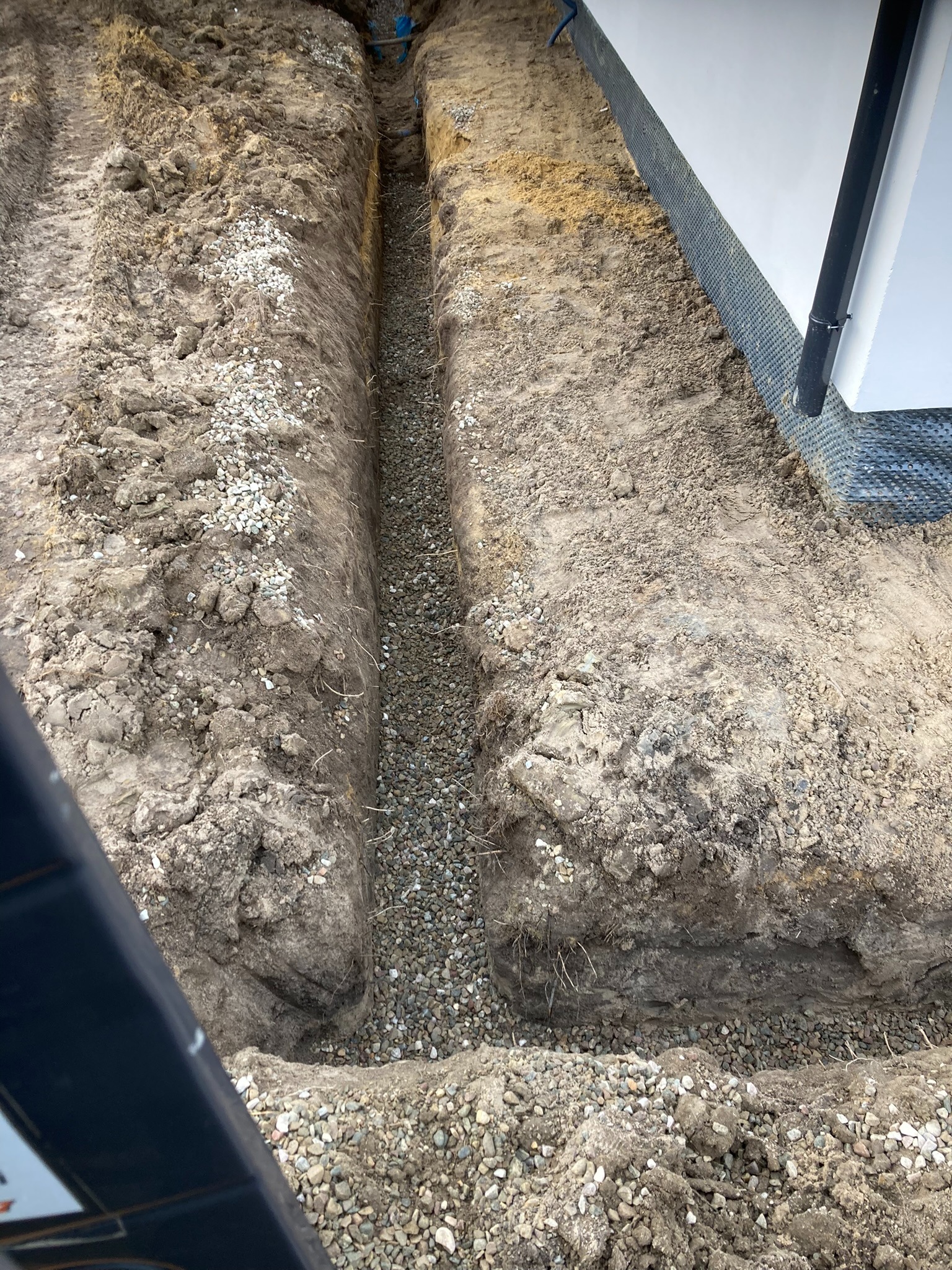 Wykopany wąski rów z podsypką żwirową wzdłuż ściany budynku z widoczną folią kubełkową i rurami. Ziemia wokół wykopu.