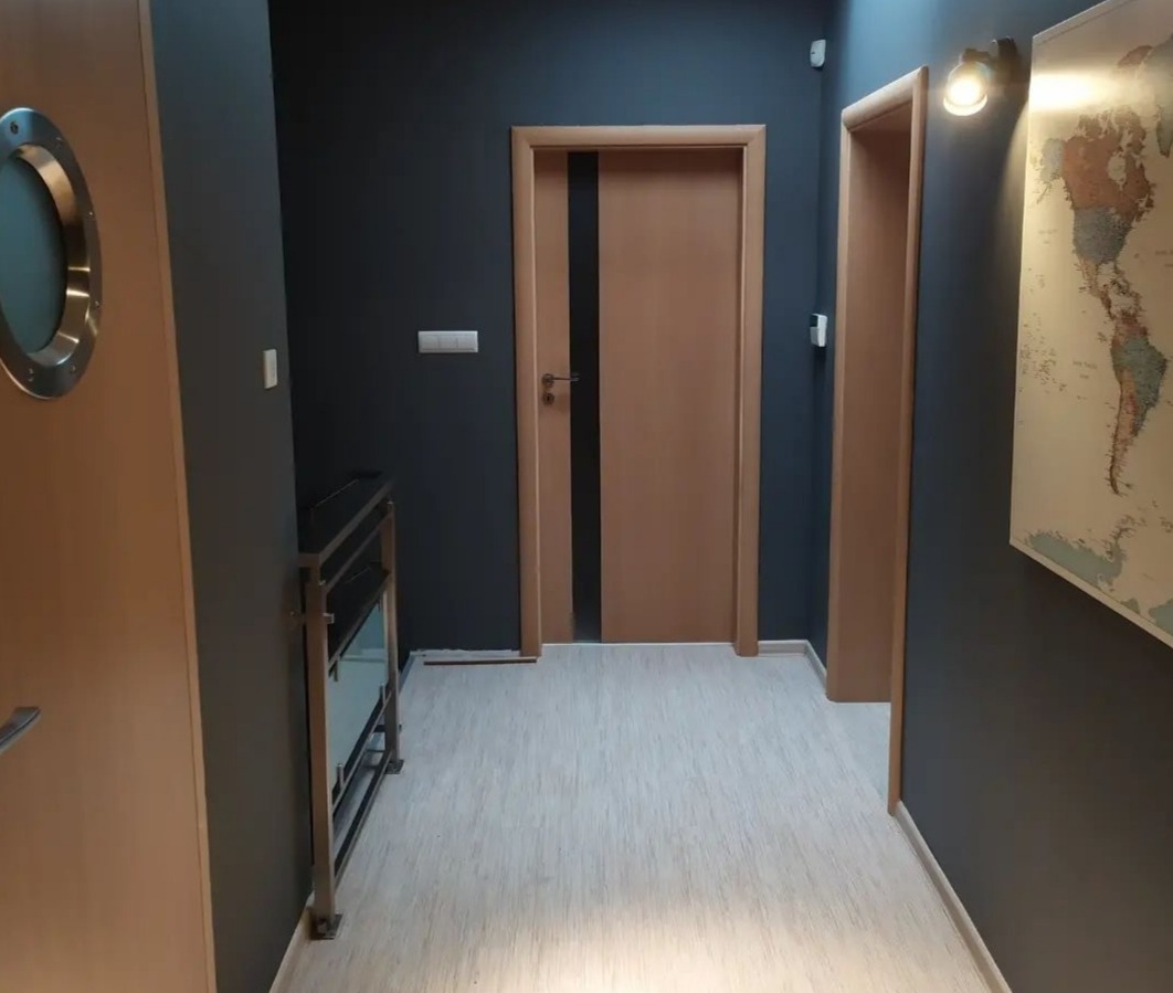 Korytarz w odcieniach szarości z drewnianymi drzwiami, okrągłym oknem w stylu bulaja i mapą świata na ścianie.