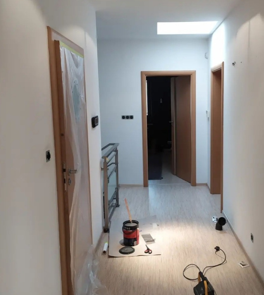 Korytarz w trakcie odświeżania, z zabezpieczonymi folią drzwiami, widoczne narzędzia malarskie, pędzel w puszce z farbą oraz lampa warsztatowa na podłodze.