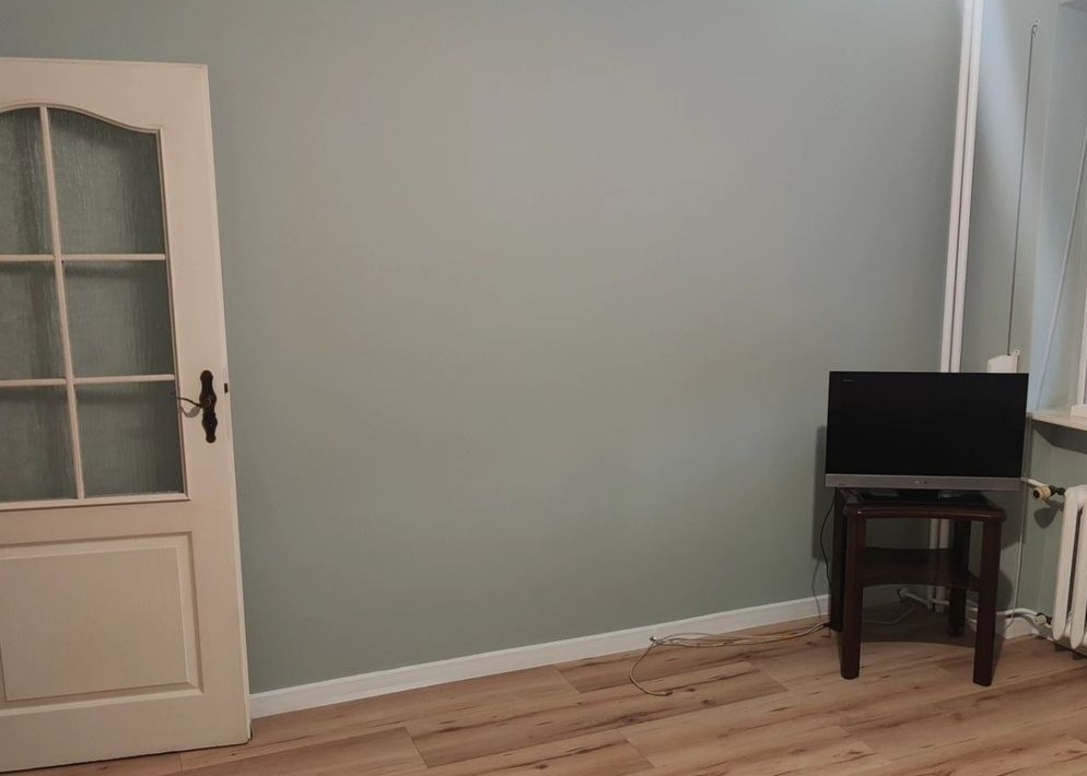 Ściana w odcieniach szarości w pokoju po świeżym pomalowaniu, widoczna biała listwa przypodłogowa, drewniana podłoga, drzwi z szybkami po lewej stronie i telewizor na stoliku po prawej.