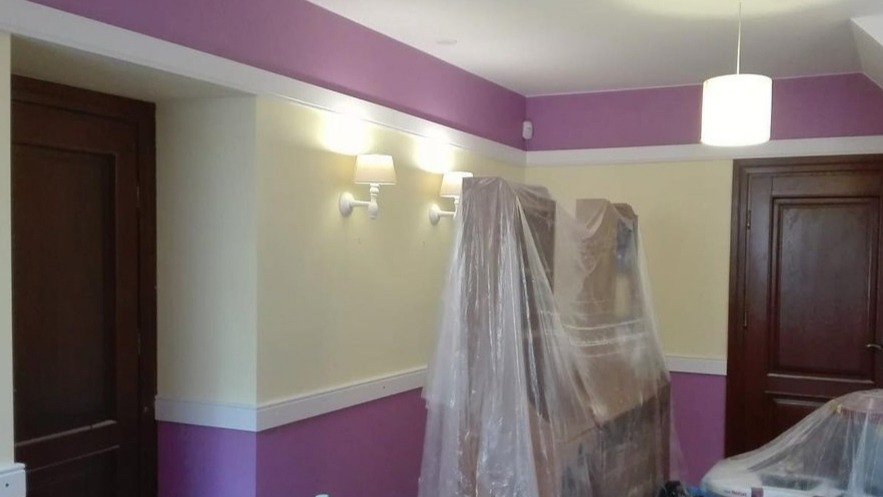 Pokój w trakcie odświeżania: ściany w dwóch odcieniach beżu i fioletu, zabezpieczone meble folią malarską, widoczne lampy ścienne i sufitowa.