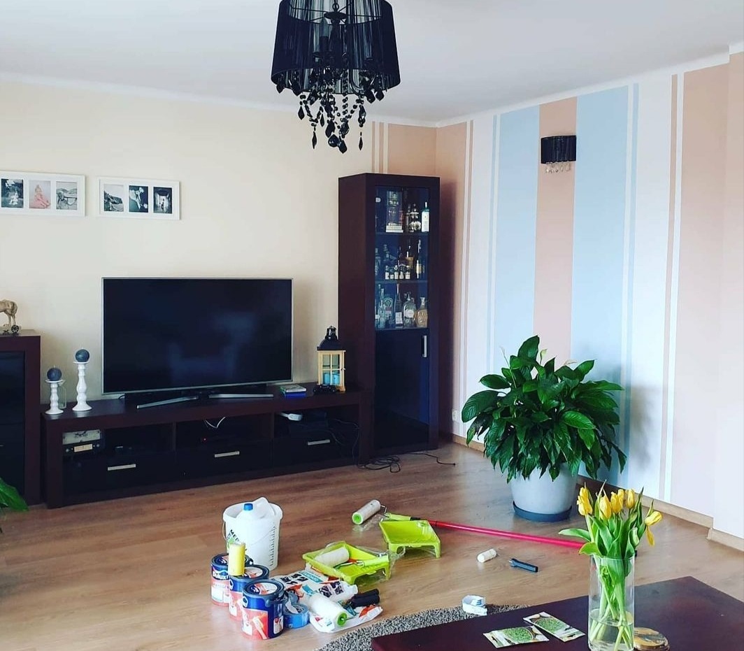Pokój dzienny z pomalowaną ścianą w pionowe pasy w odcieniach różu i błękitu, na podłodze rozłożone narzędzia malarskie, w tle telewizor i czarny żyrandol.