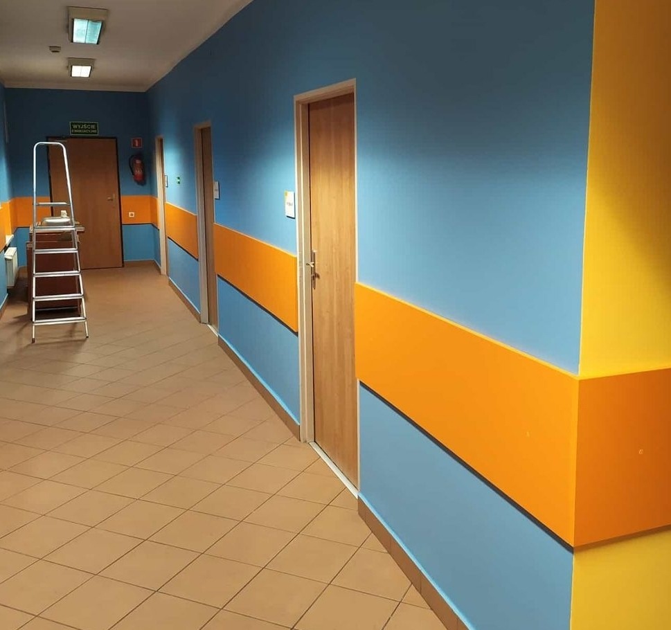 Korytarz po odświeżeniu: ściany pomalowane na niebiesko z poziomym pasem w kolorze pomarańczowym, narożnik w kolorze żółtym, widoczna drabina malarska.