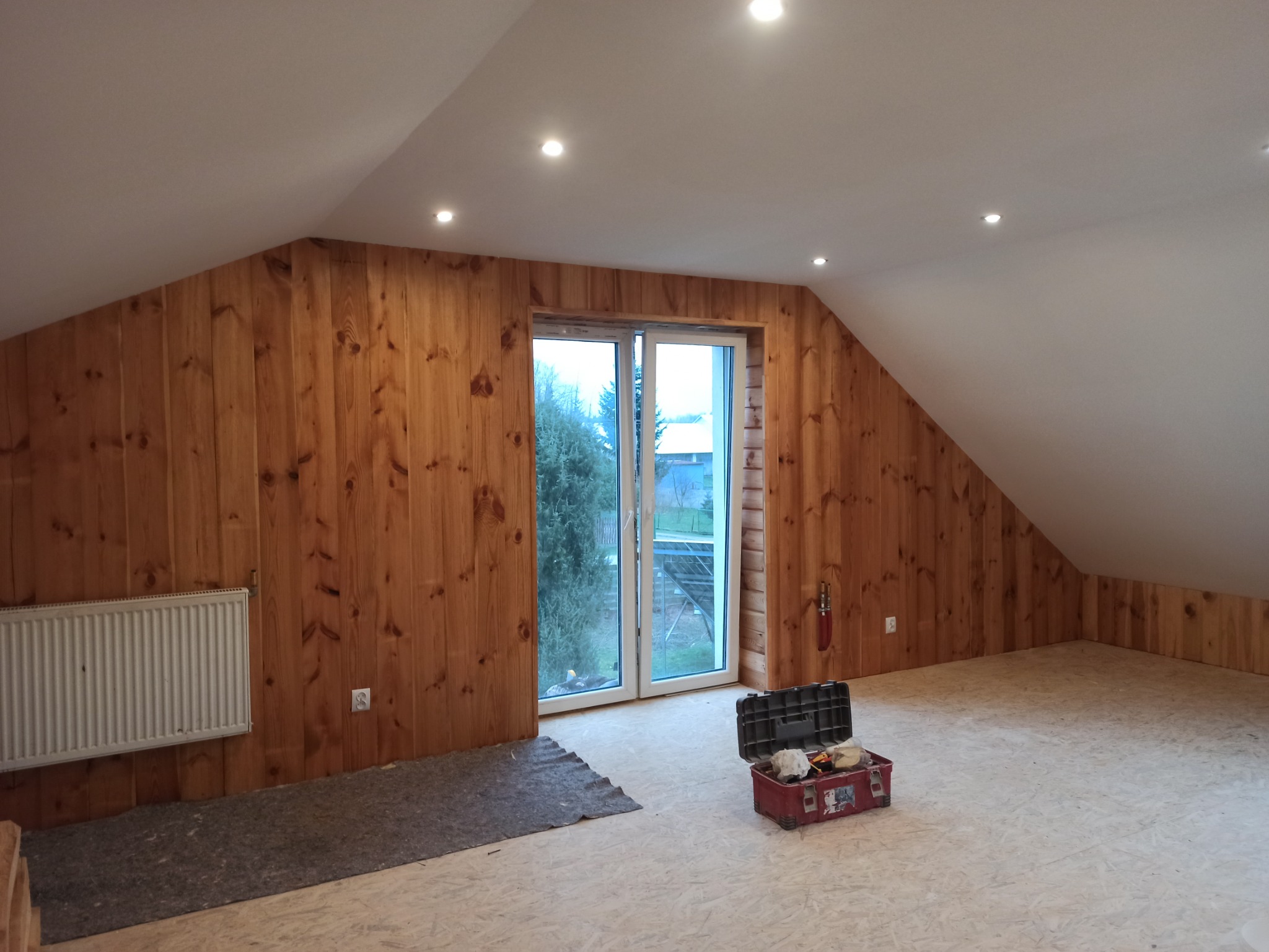 Poddasze po remoncie, ściany wyłożone drewnianą boazerią, widoczne okno balkonowe, grzejnik, skrzynka z narzędziami i fragment wykładziny ochronnej na podłodze.
