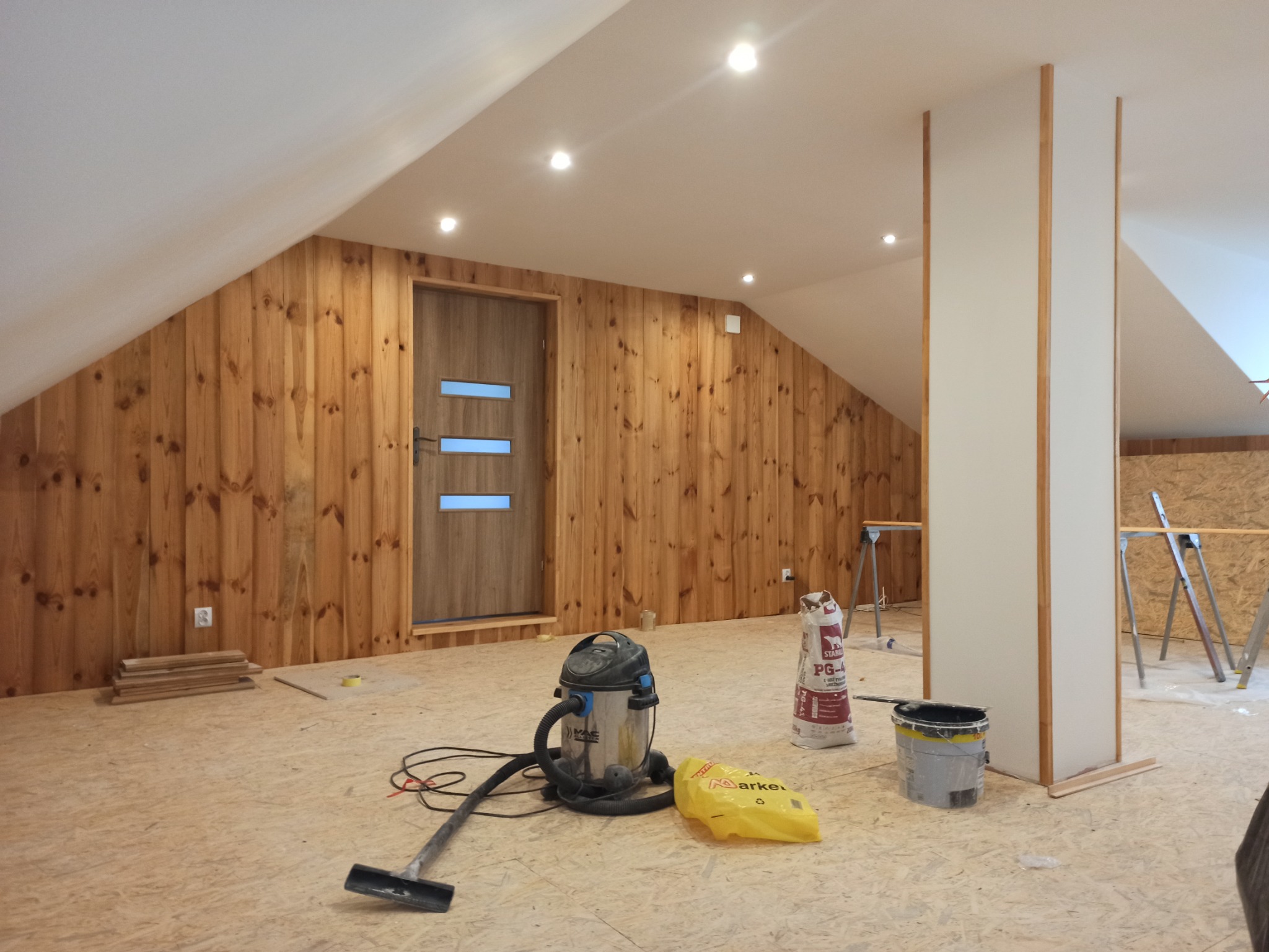Poddasze w trakcie wykańczania: boazeria z jasnego drewna, dwoje drzwi z podłużnymi okienkami, odkurzacz przemysłowy, worek z zaprawą, wiadro z farbą oraz fragmenty płyt OSB na podłodze.