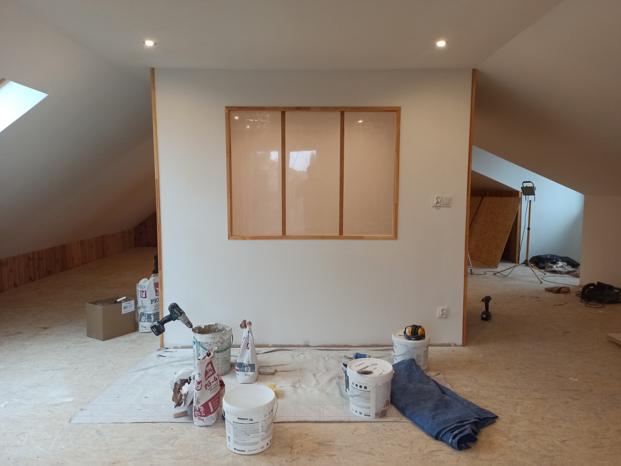 Poddasze w trakcie prac wykończeniowych: biała ściana z drewnianą ramą okienną, zakryta folią, widoczne wiadra z farbą, gładzią i narzędzia na zabezpieczonej podłodze.