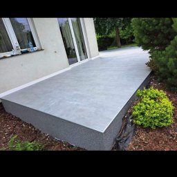 Remont - Wykończony taras z szarą, gładką powierzchnią, obłożony drobnym kamieniem, przylegający do białej ściany budynku z oknami i drzwiami tarasowymi, otoczony zielenią.