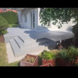 Szary taras z płytkami ceramicznymi i schodami, widok z poziomu ogrodu, donice z ziołami w pierwszym planie, budynek w tle.