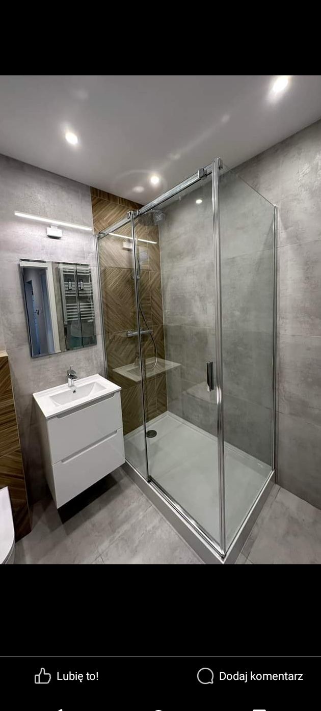 Nowoczesna łazienka z kabiną prysznicową z przesuwnymi drzwiami, białą umywalką z szafką i lustrem z oświetleniem LED, ściany wykończone szarymi płytkami imitującymi beton i panelami drewnopodobnymi.